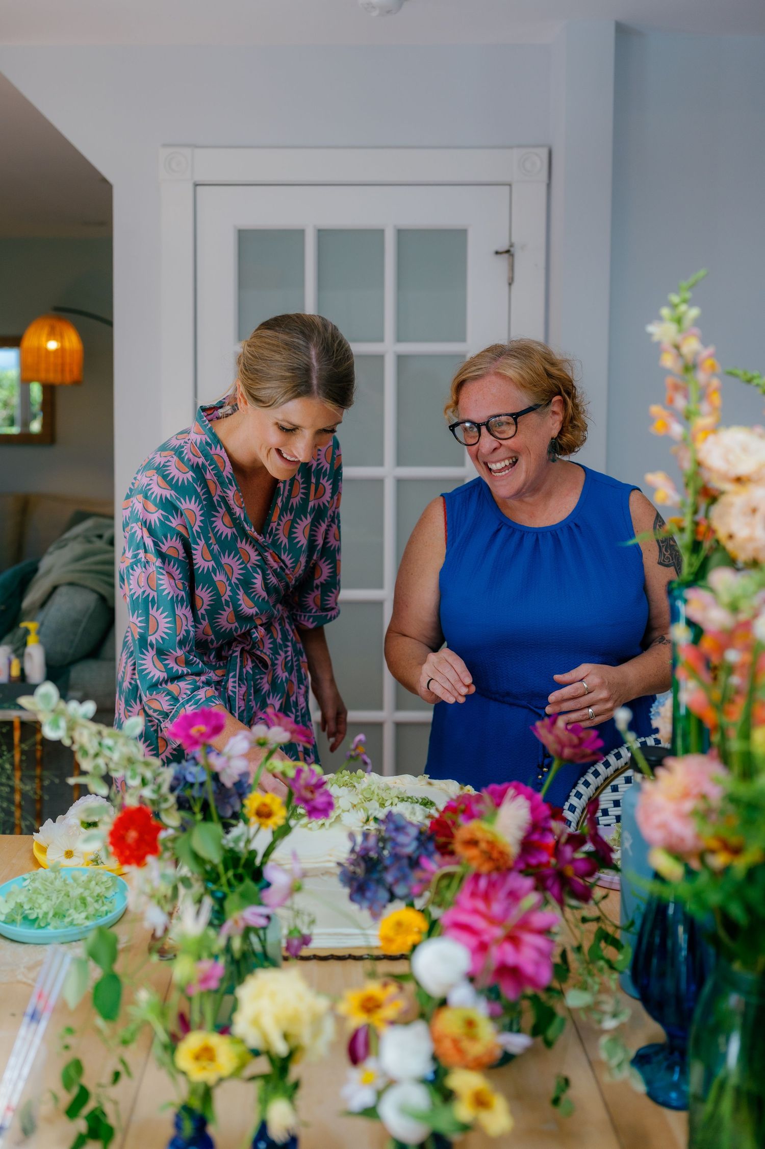 Two women laughing together arranging colorful fresh flowers on a table indoors at a bright floral workshop.