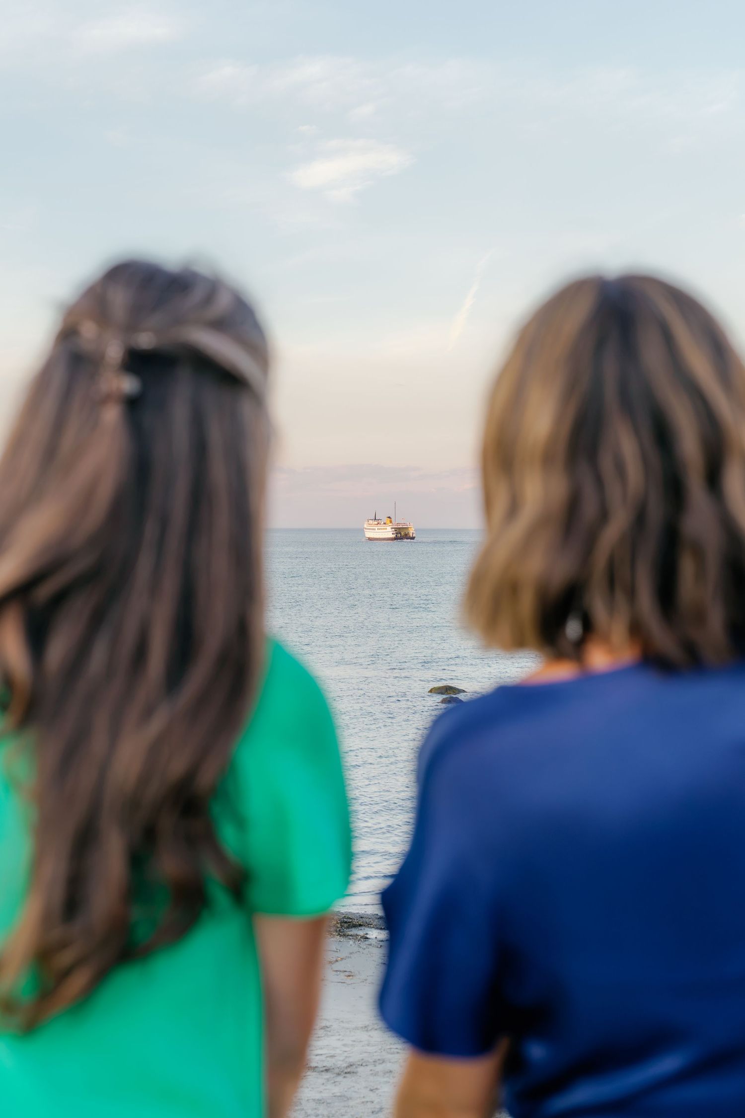 Two people with long hair view a ship on the calm ocean horizon from a beach at dusk.