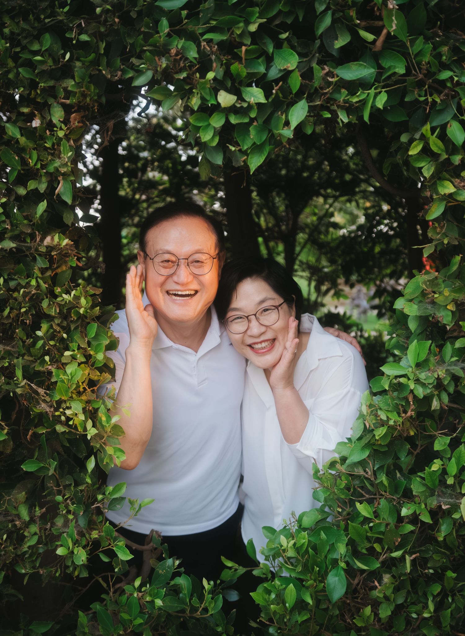 Two smiling people wearing glasses and white shirts pose together among lush green leafy bushes outdoors.