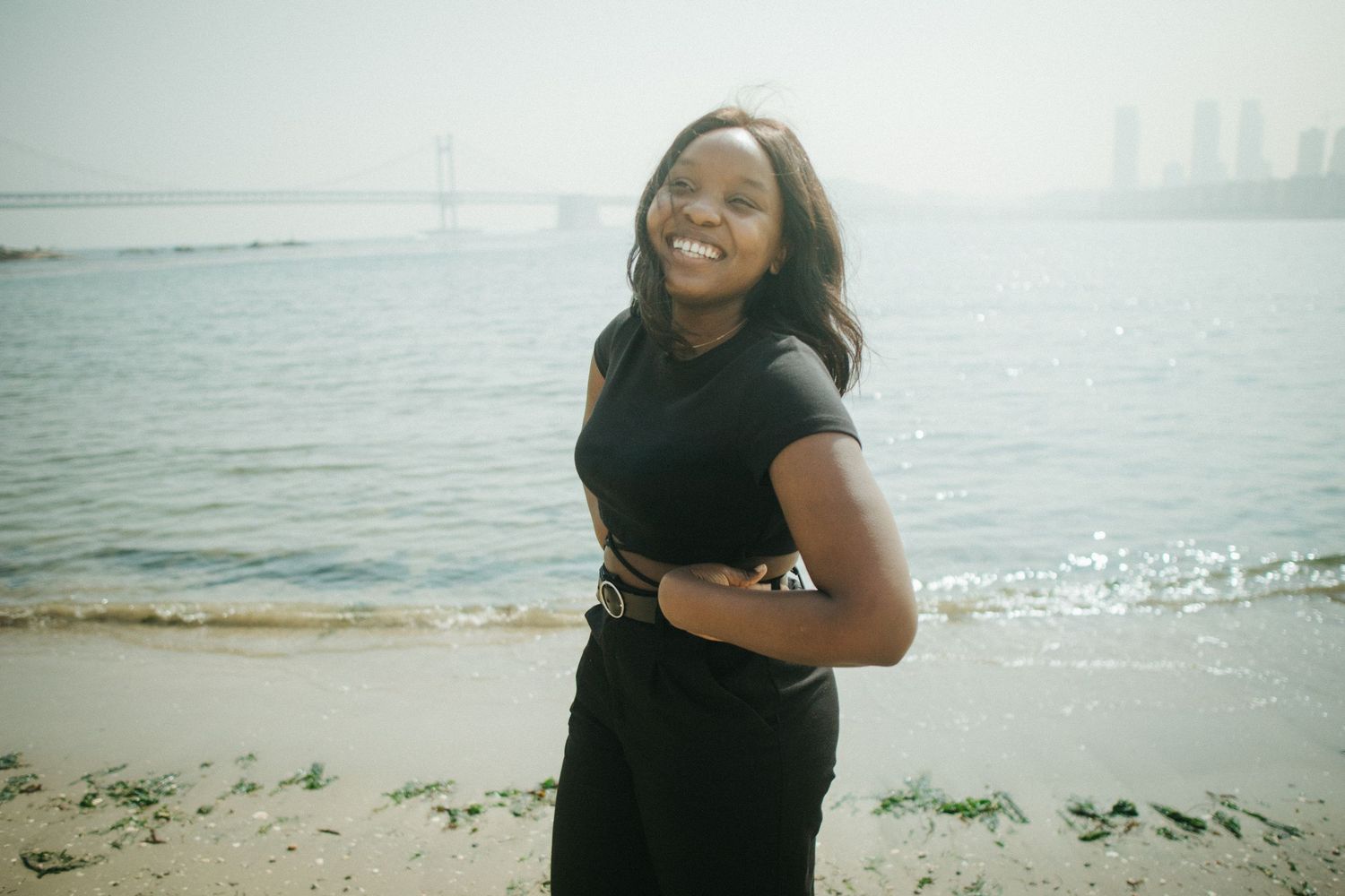 Smiling woman in black outfit standing on a sunny beach with a bridge and city skyline behind.
