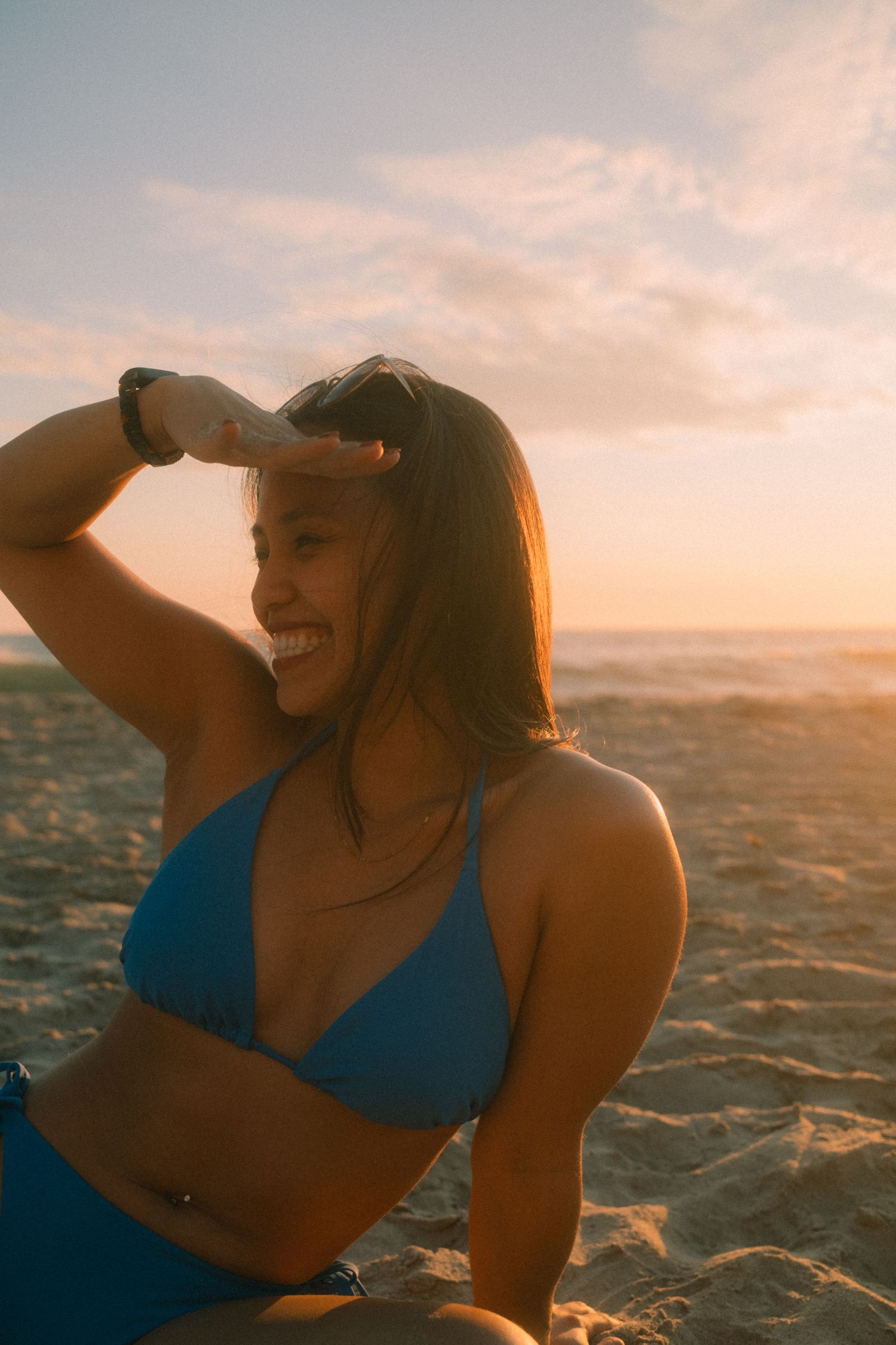 Smiling woman in blue bikini on sandy beach shading eyes during sunset.