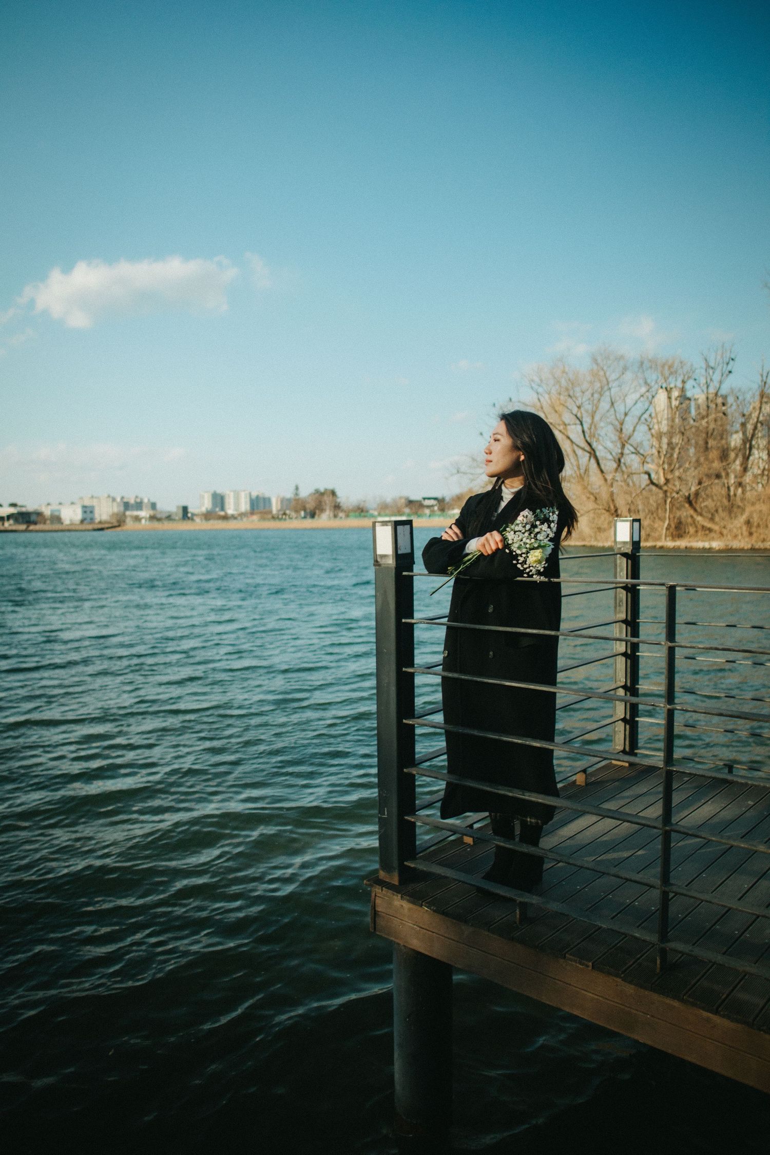 Woman in black embroidered coat standing on wooden pier overlooking blue lake under clear sky.