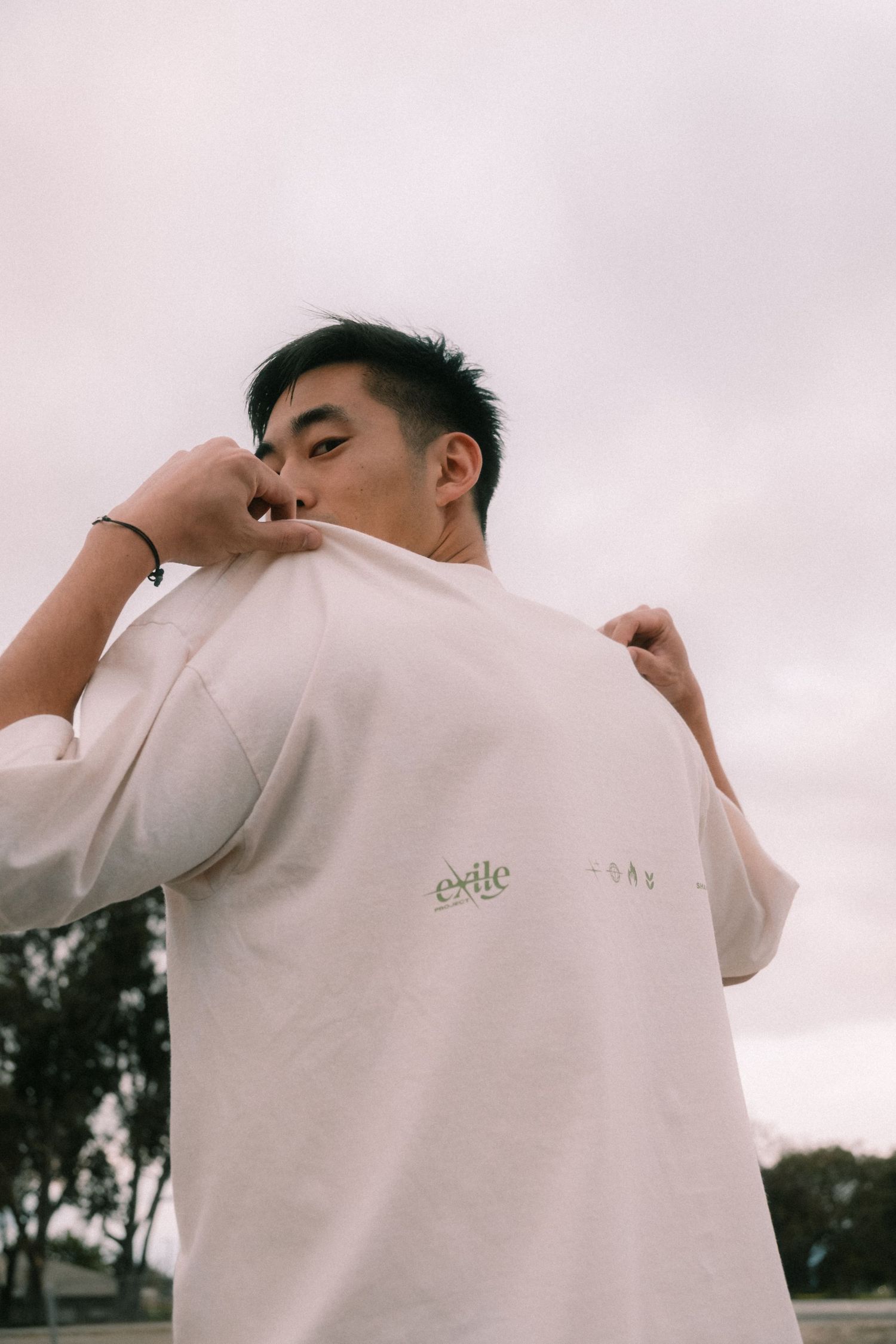 Young man in oversized cream sweatshirt with Exile logo pulling up collar against cloudy sky.