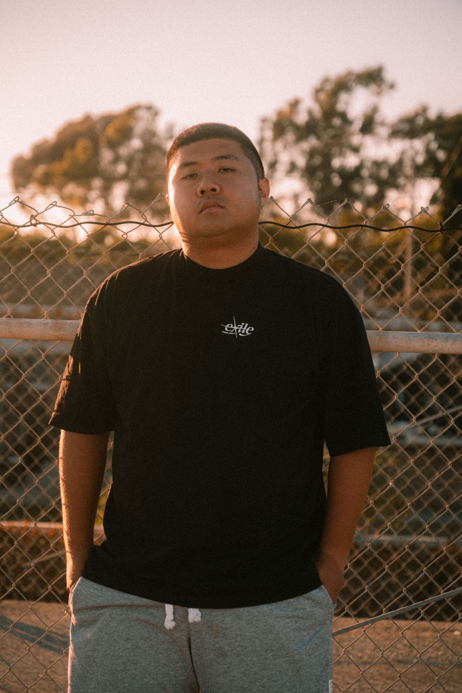 Man in oversized black graphic tee standing by chain-link fence at golden hour.