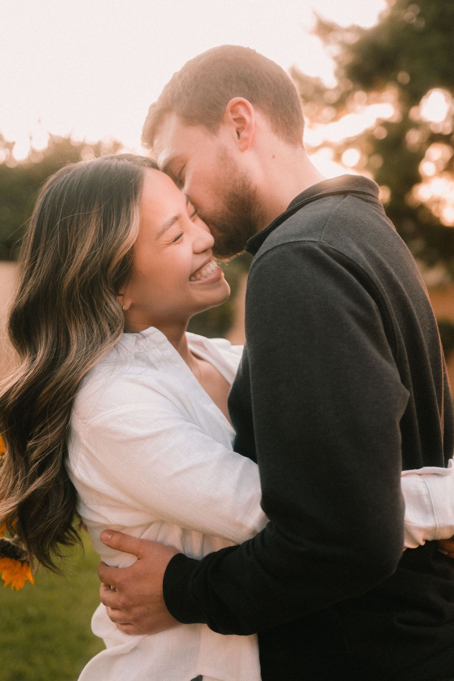 A joyful couple embraces at golden hour, the smiling woman in white hugging a man in black as sunlight glows behind them.