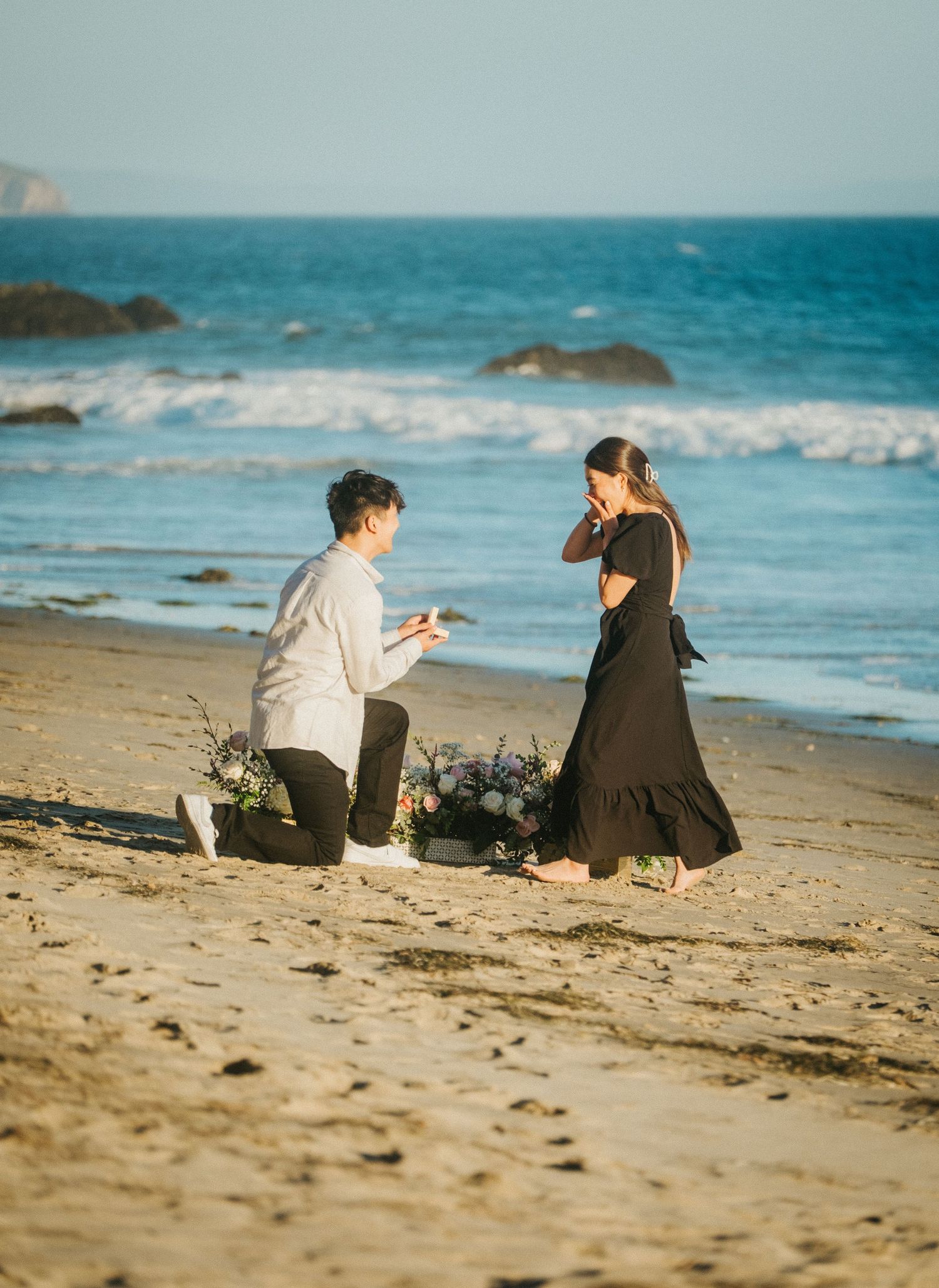 Man proposes to woman on a sandy beach with flowers, ocean waves in the background, as she covers her mouth in surprise.