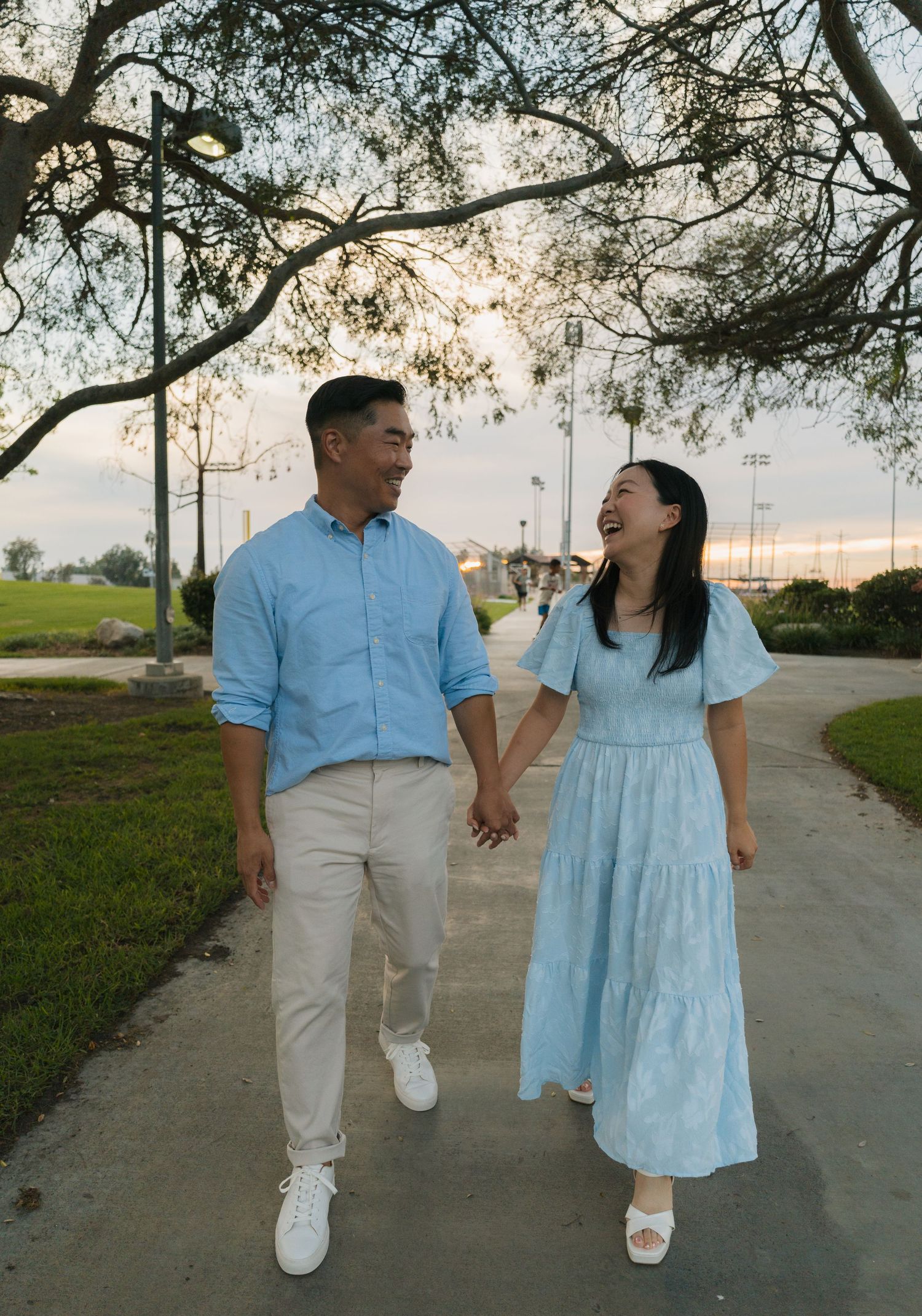 A smiling couple holds hands and walks together on a park path at sunset, dressed in coordinating light blue outfits.