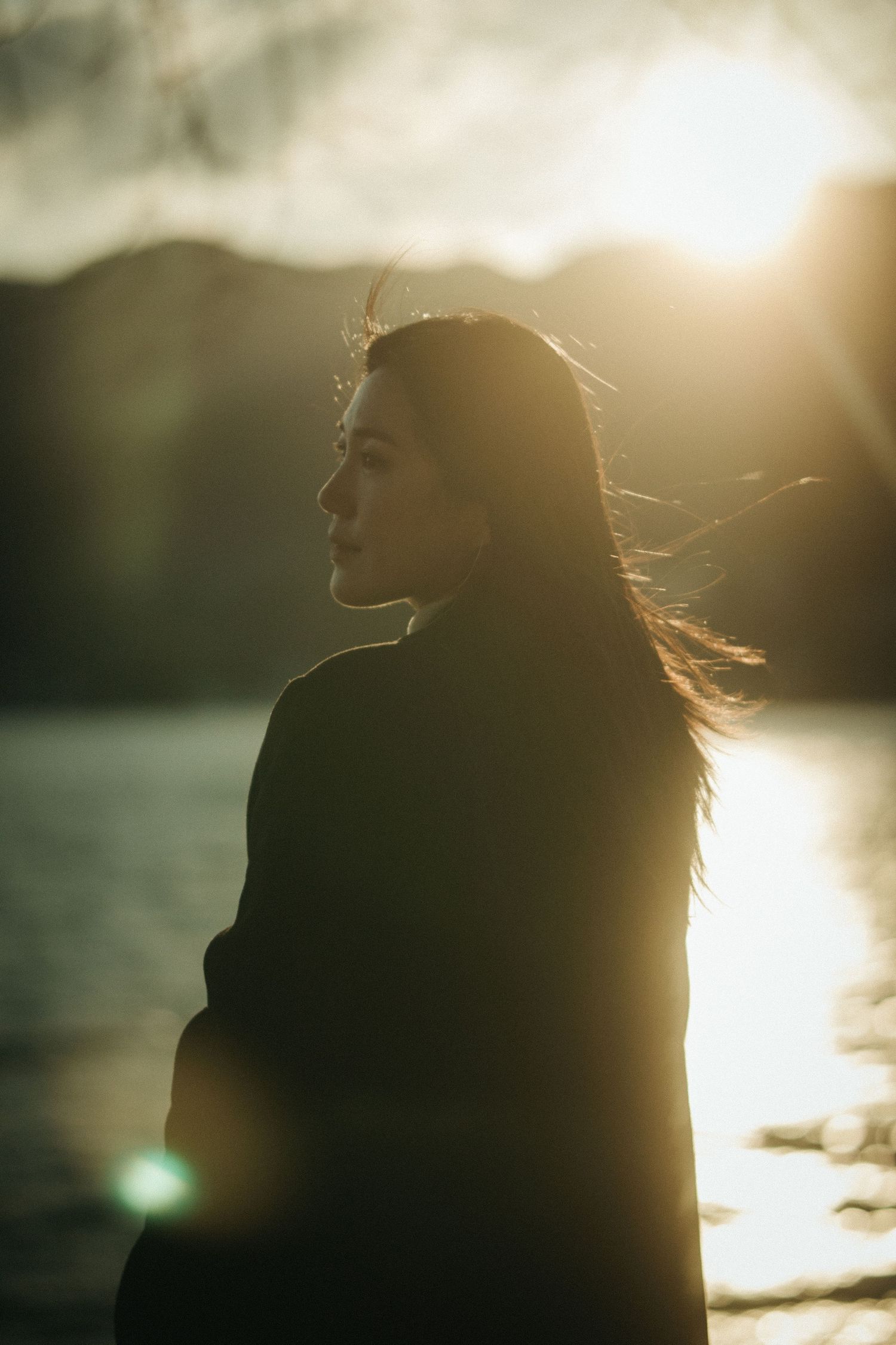 Serene side-profile silhouette of woman with windswept hair in warm golden sunset glow by lake.