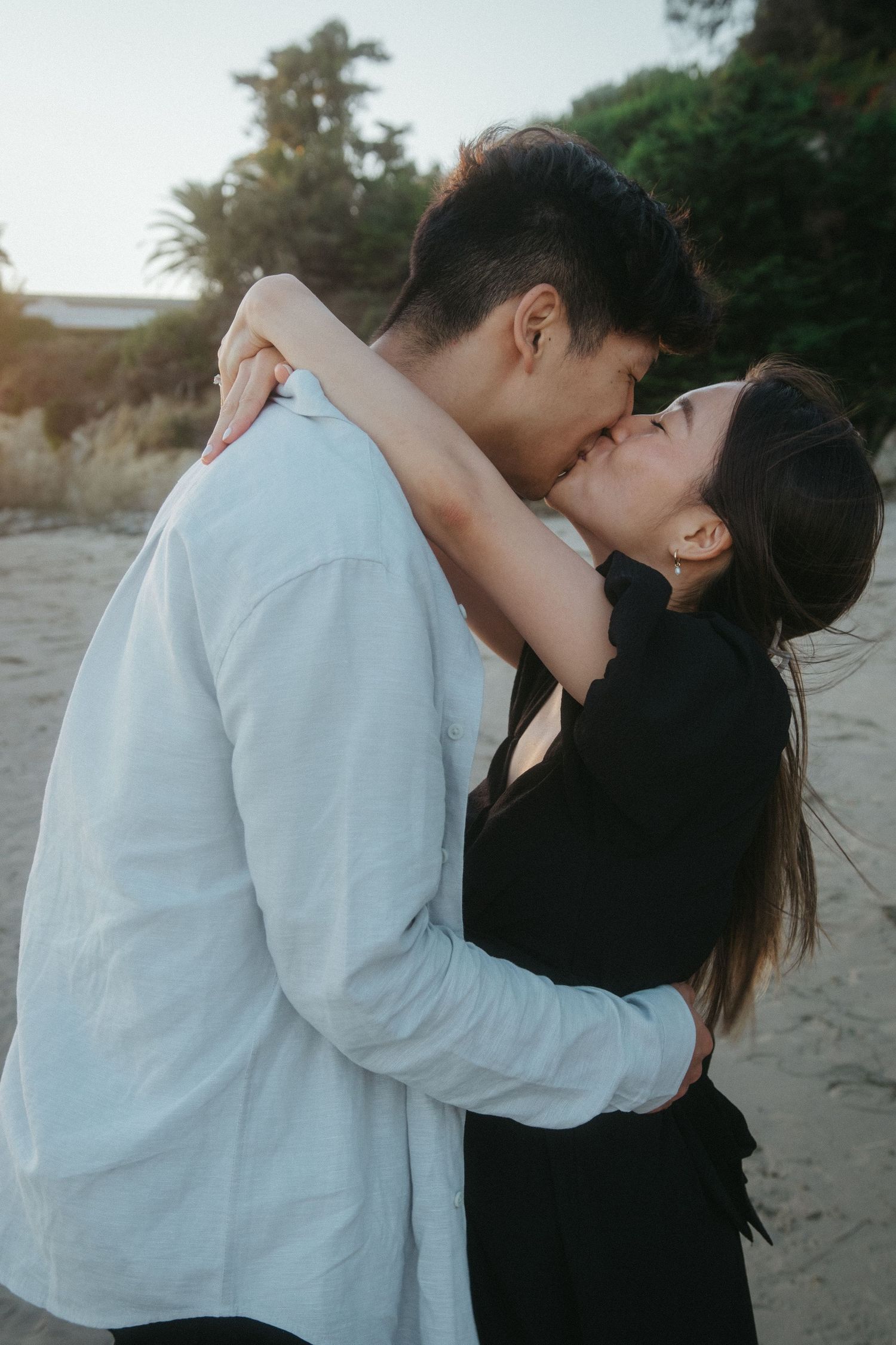 Newly engaged Asian couple sharing a passionate kiss on the beach at golden hour.
