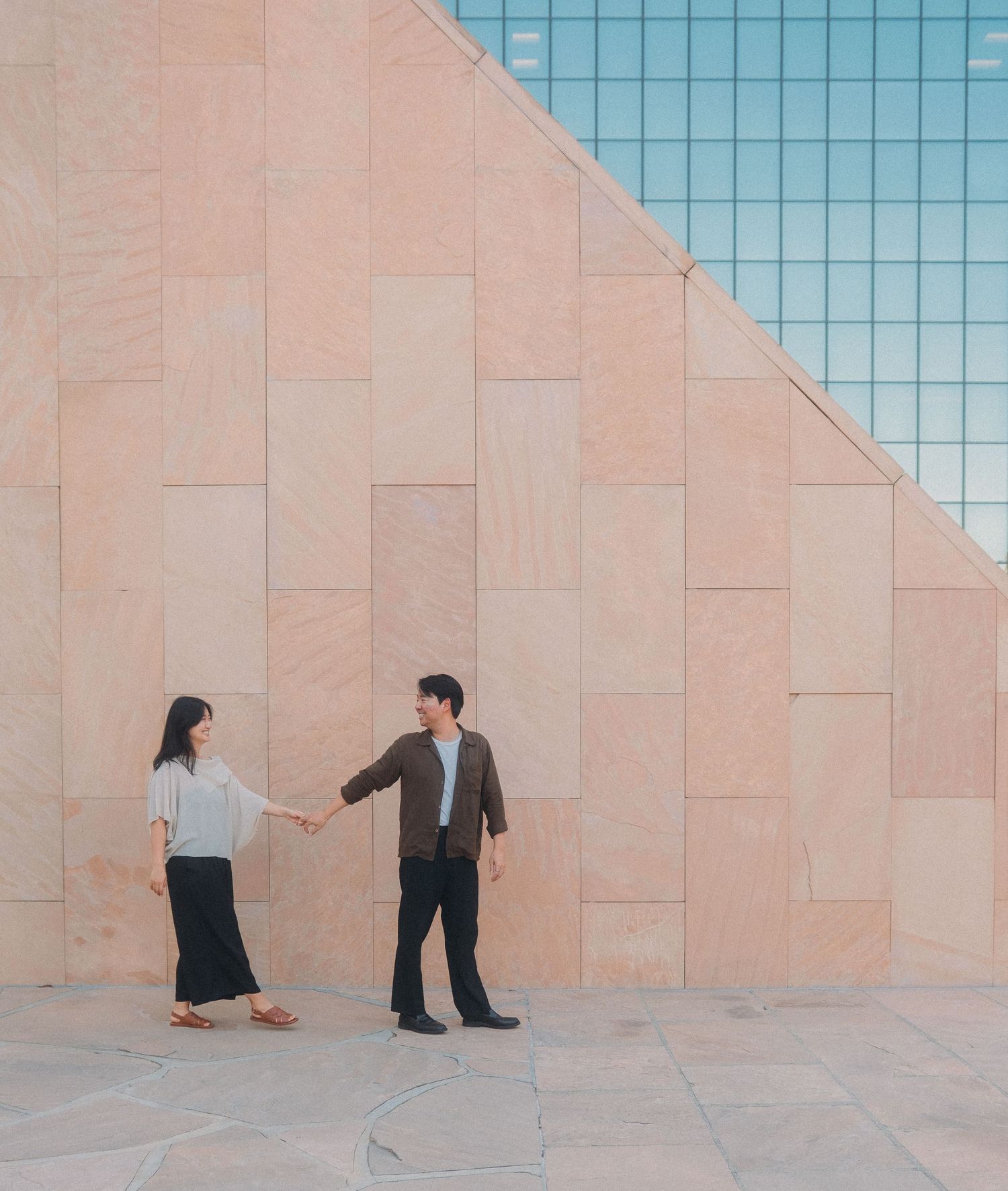 Asian couple holding hands and facing each other against geometric pink stone wall with glass building.