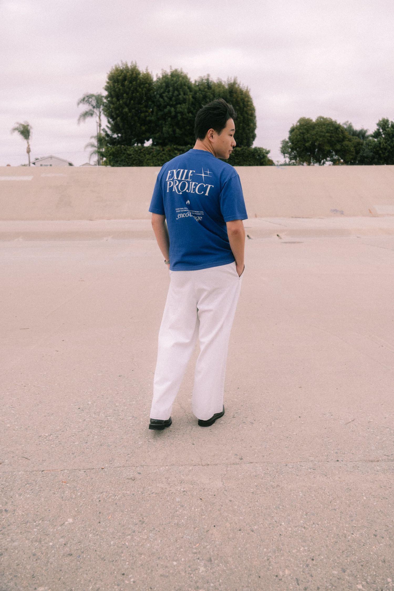 Man in blue Exile Project graphic tee and white pants standing on open pavement outdoors.