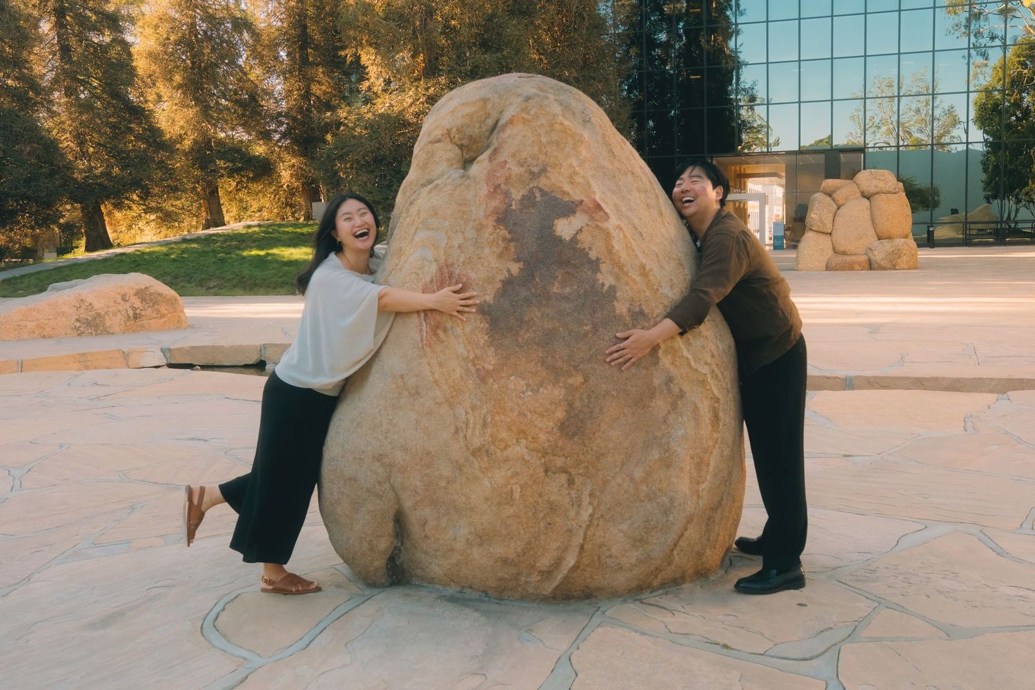 Laughing Asian couple playfully posing with a large boulder sculpture in an outdoor park setting.