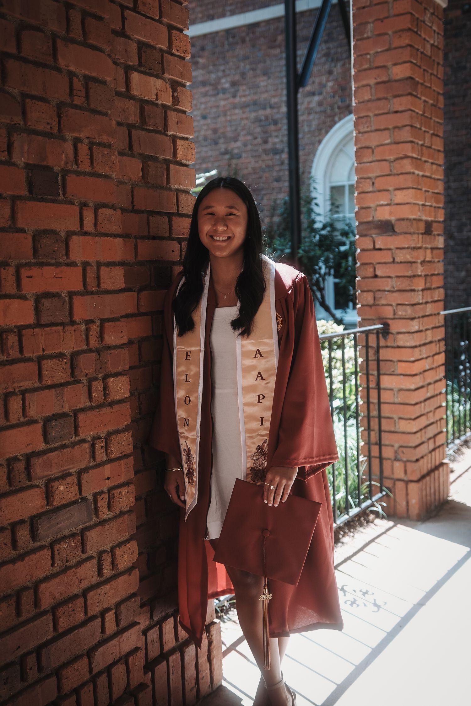 Smiling graduate in maroon gown with AAPI stole leans against brick wall.