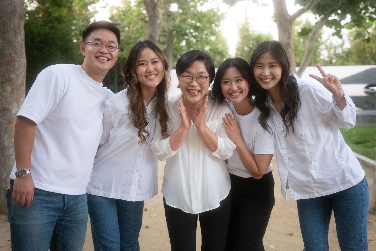 Five smiling young Asian adults in white shirts pose together outdoors among trees in a cheerful group photo.