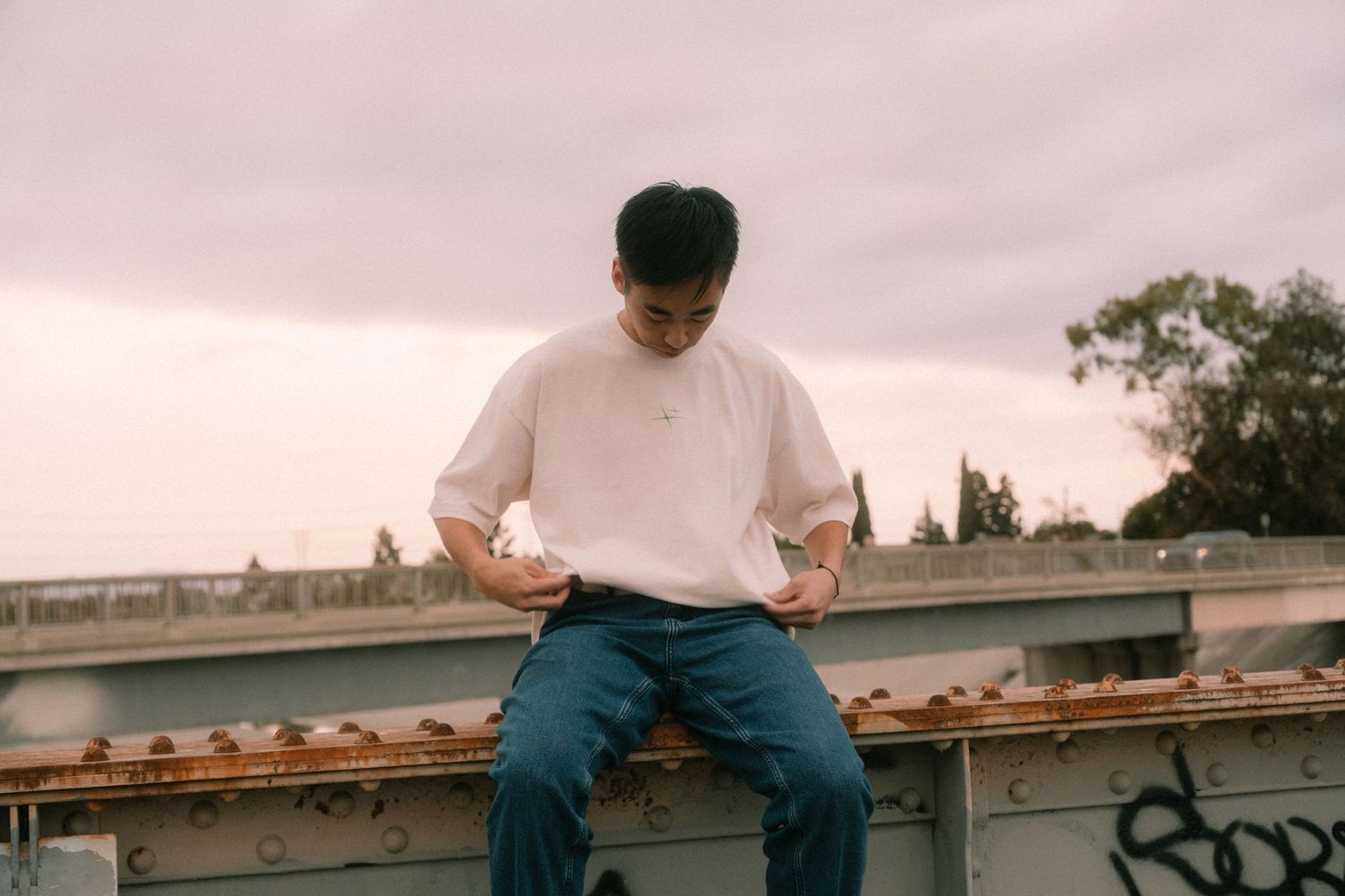 Young man in white t-shirt and blue jeans sitting on a rusty metal structure outdoors under cloudy sky.