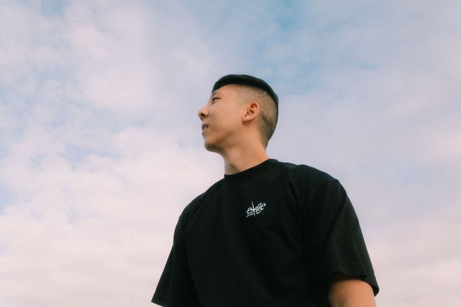 Young Asian man in black t-shirt gazing upward against a cloudy blue sky, shot from low angle.