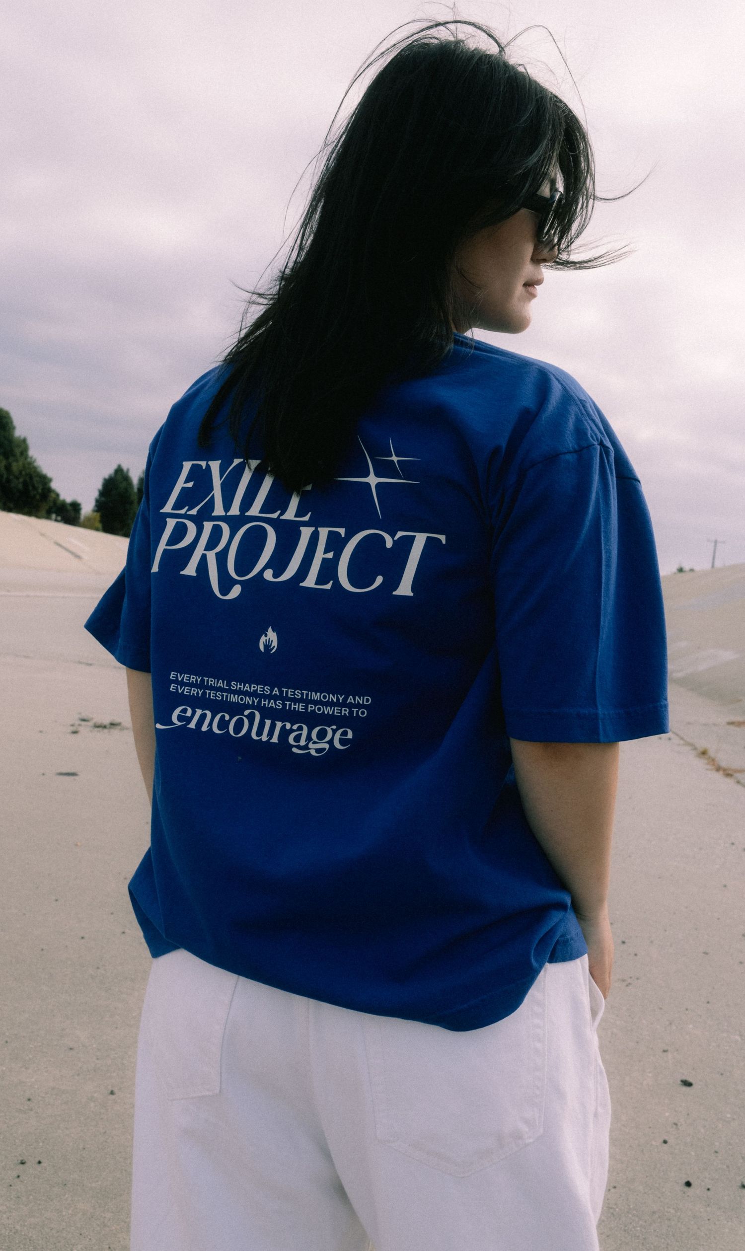 Woman showing back of blue Exile Project encourage graphic tee on sandy beach.