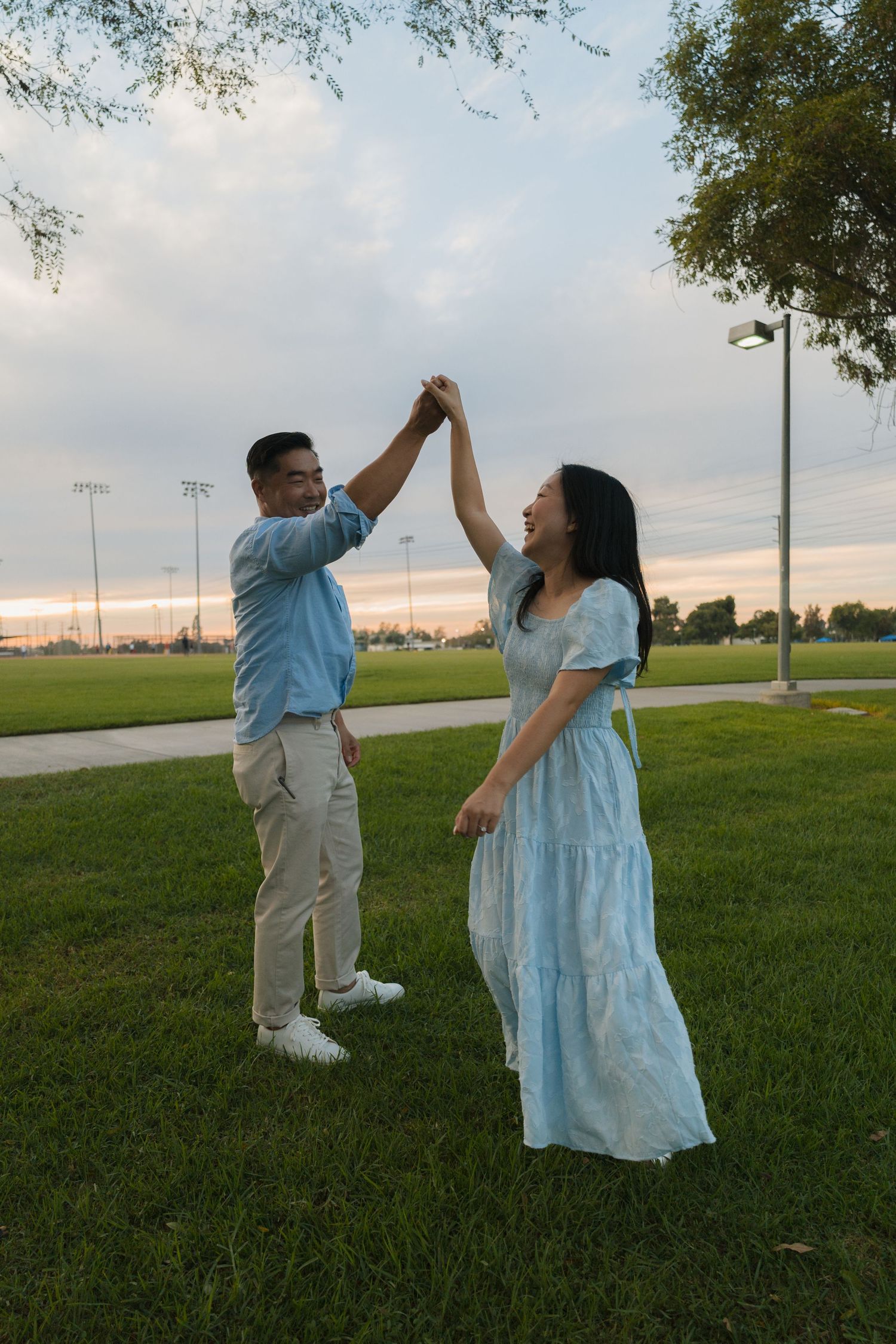 Happy couple dancing together on green grass in park at sunset, man spinning woman in blue dress.