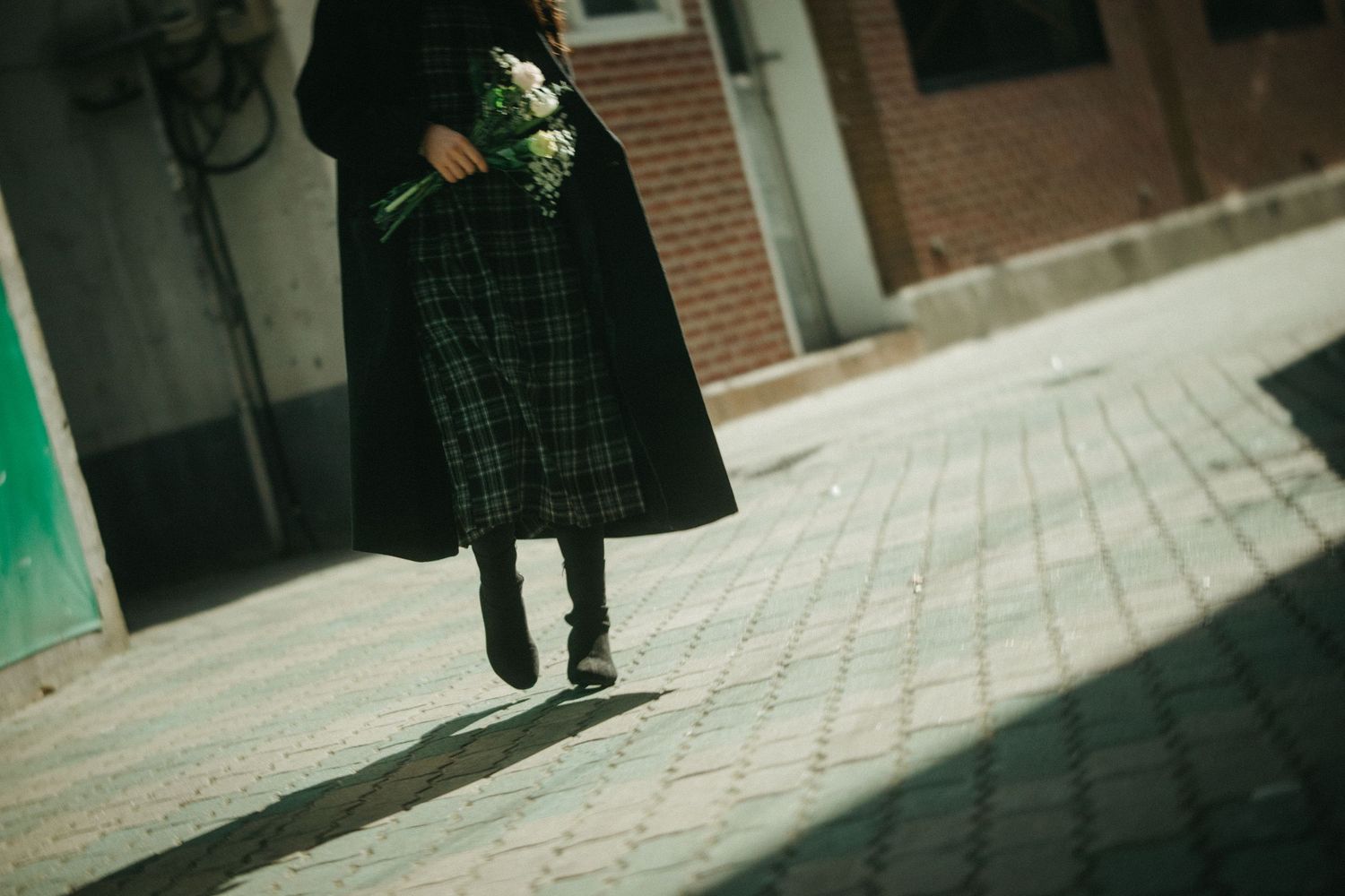 Close-up of woman in plaid skirt and black coat holding white flowers on sunlit cobblestone path.