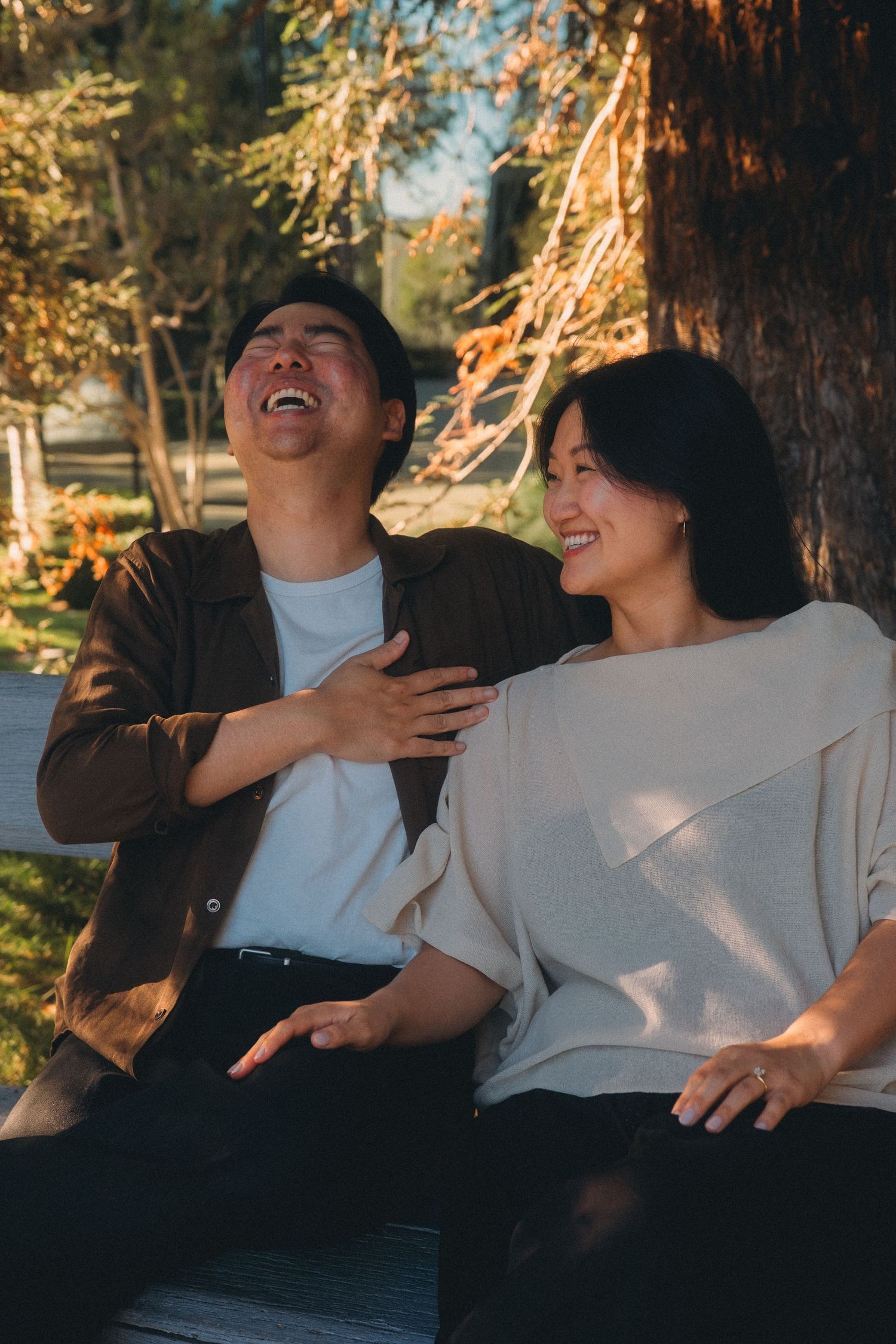 Asian couple laughing and smiling together on a bench in a sunlit outdoor park setting.