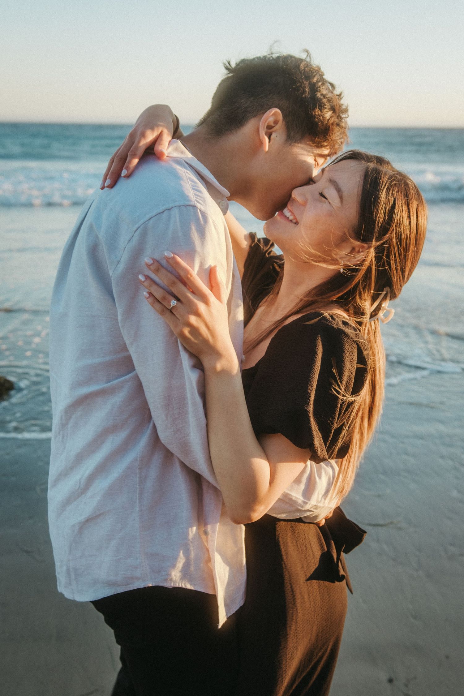 Romantic couple embracing and almost kissing on a beach at sunset with ocean waves behind them.