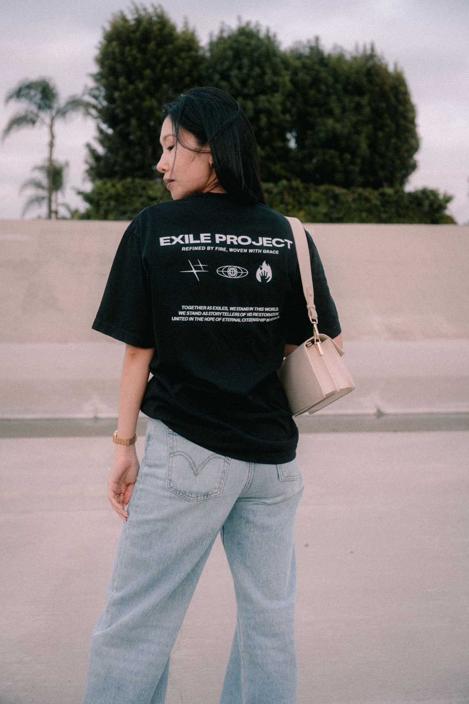 Woman wearing black Exile Project t-shirt with jeans and beige bag viewed from behind outdoors.