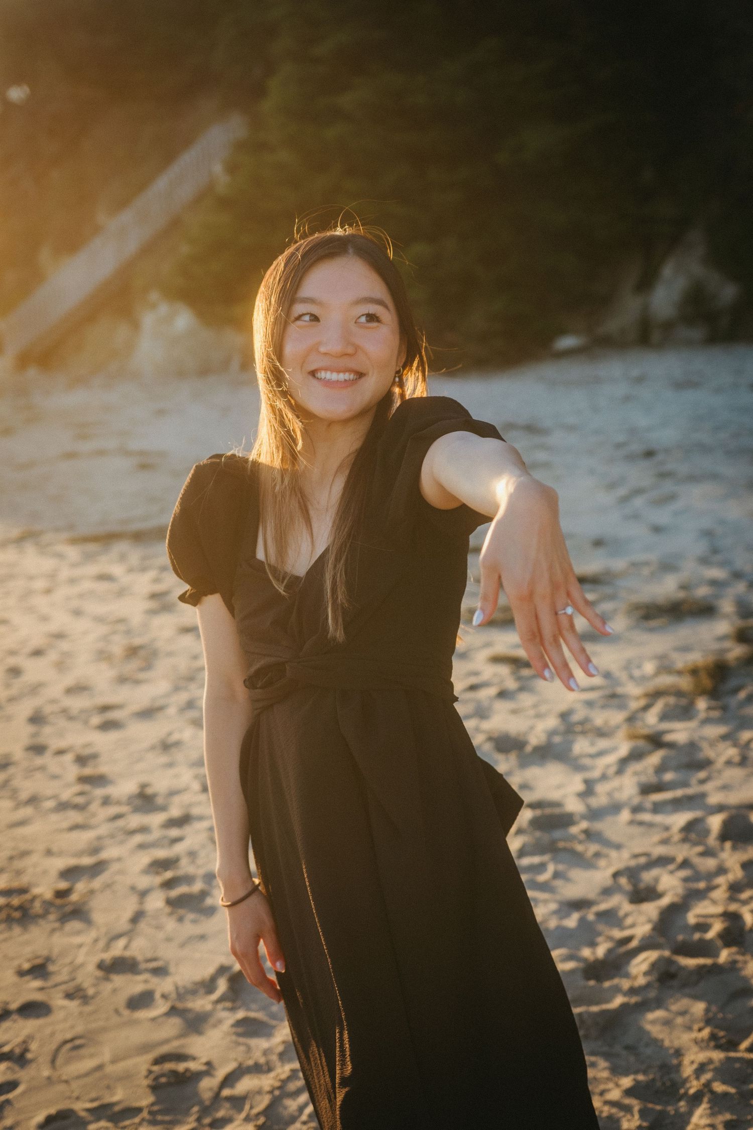 Smiling young woman in a black dress posing on a sandy beach during golden hour sunset.