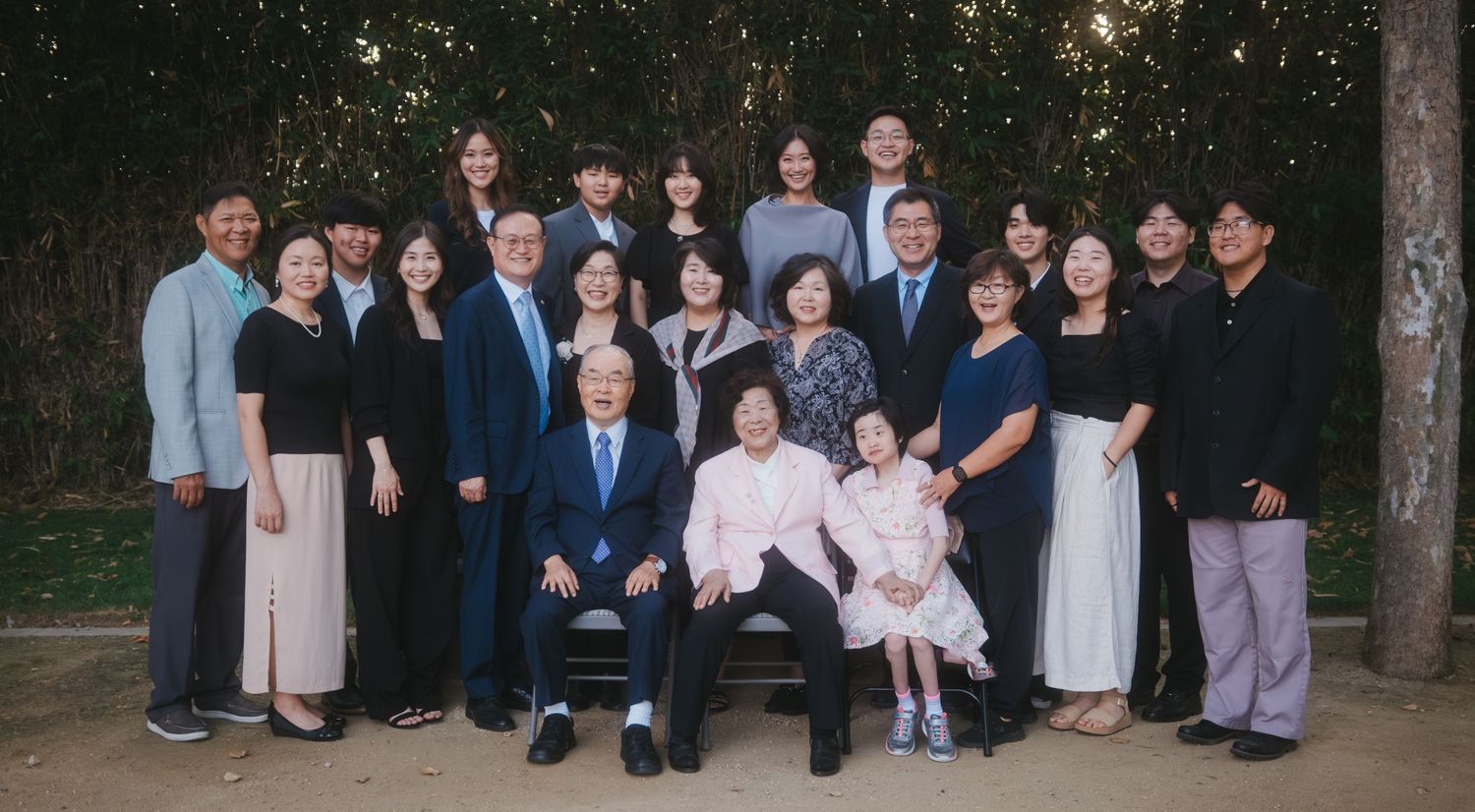 Large Asian family group portrait outdoors, dressed in formal attire, smiling together near trees.