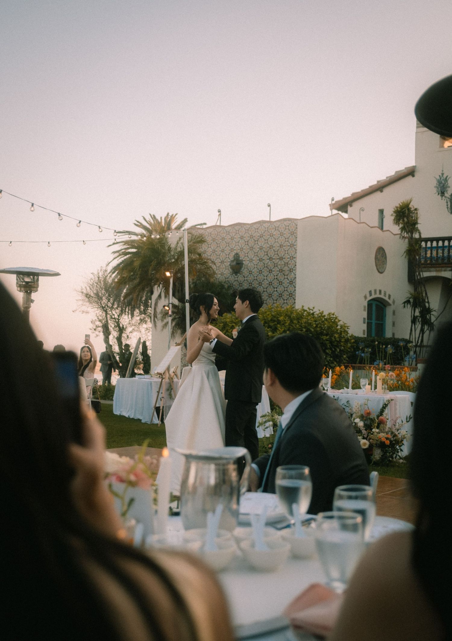 Bride and groom share first dance at outdoor wedding reception at dusk.