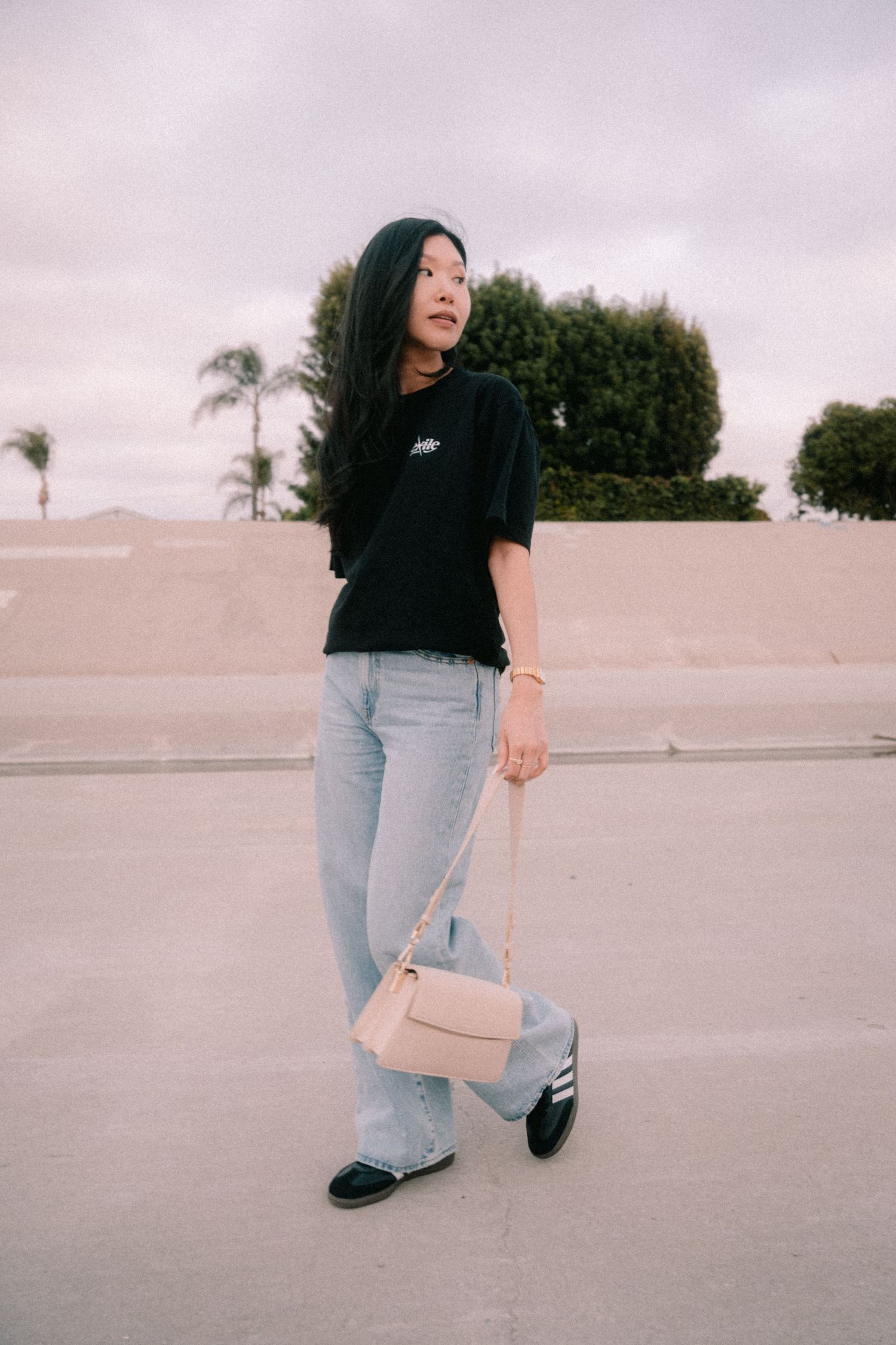 Young woman in black Exile Project tee, light jeans, holding a beige crossbody bag outdoors.