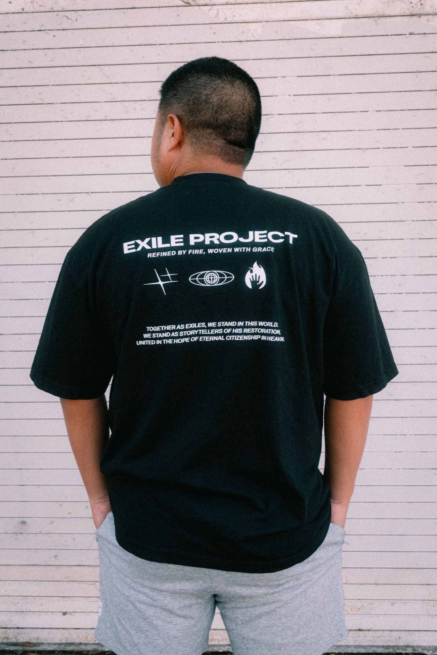 Man wearing black Exile Project graphic tee with white text, paired with gray shorts, standing against a white wall.