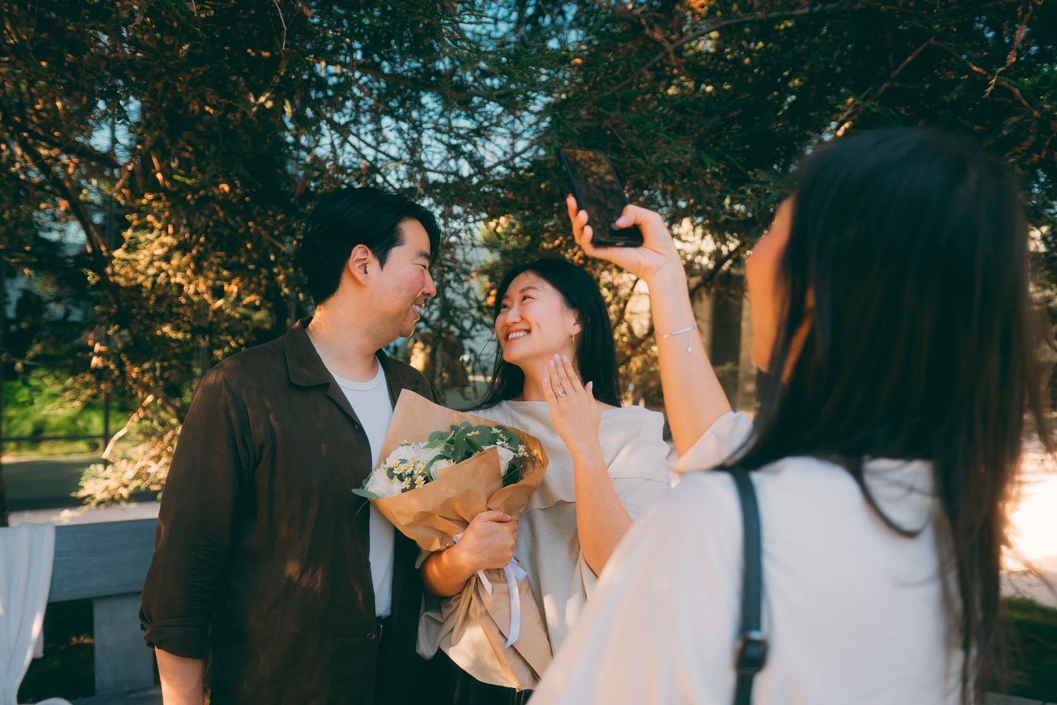 Asian couple poses for engagement photos outdoors while friend captures the moment on phone.