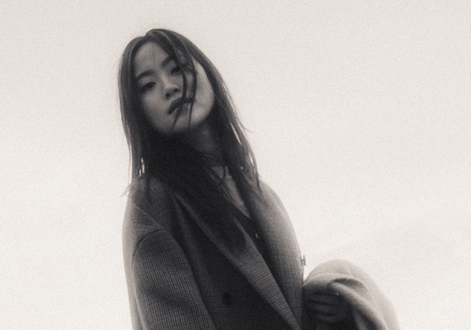 Black and white portrait of a young woman with long dark hair wearing an oversized coat outdoors.