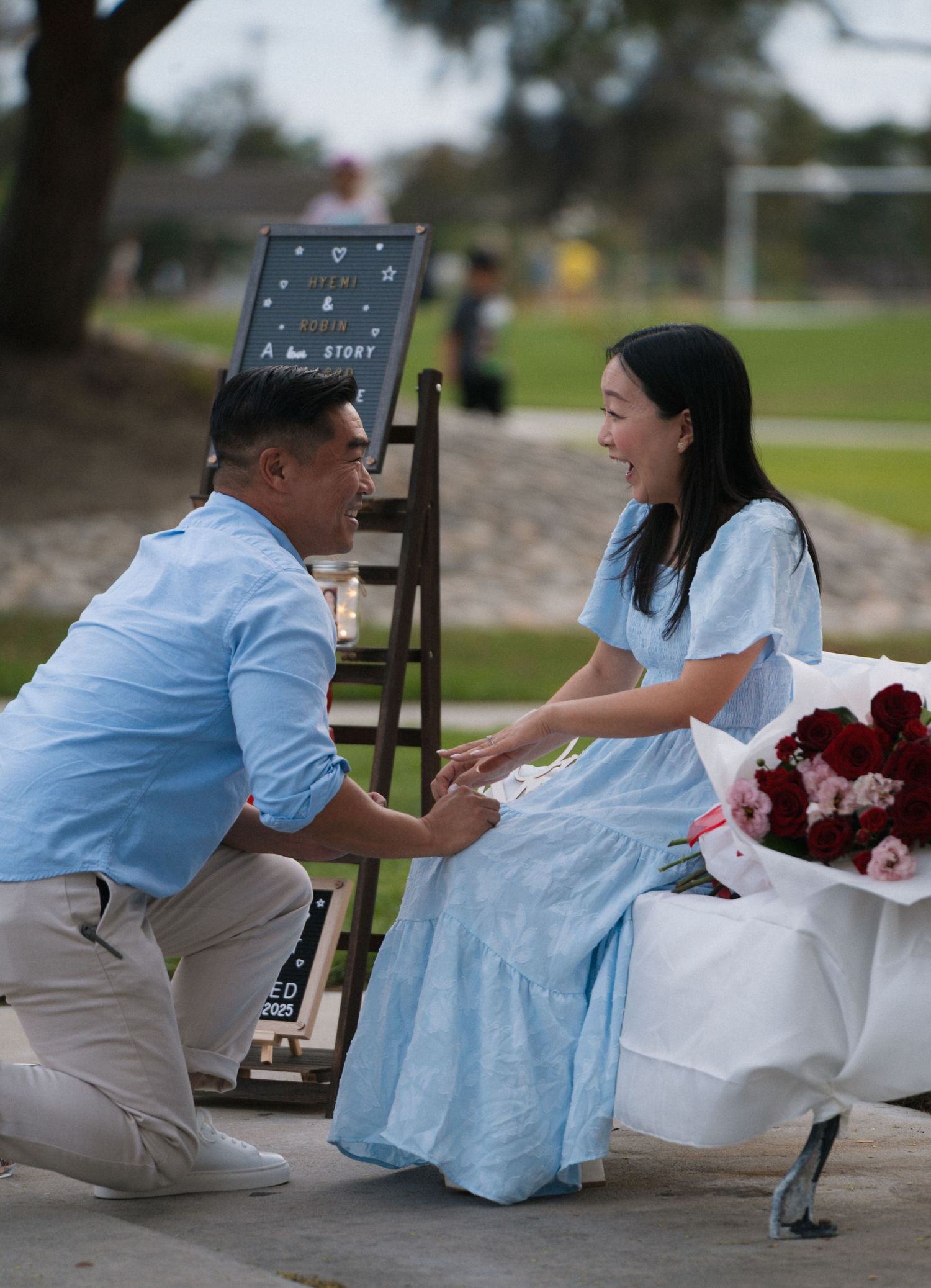A man kneels on one knee proposing to a smiling woman in a blue dress outdoors near a decorative sign and red roses.