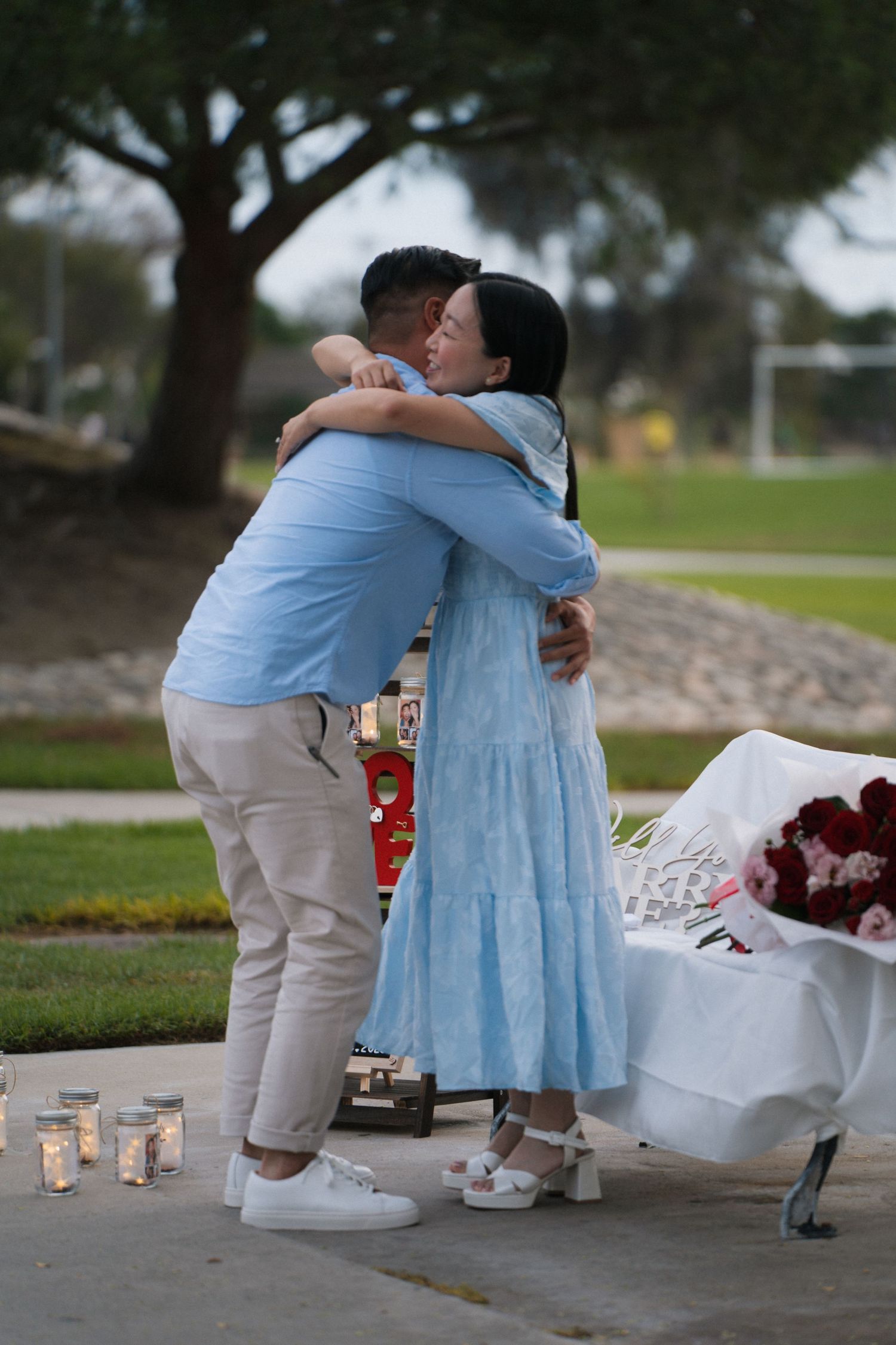 Couple in matching blue outfits sharing a romantic hug near candles and red roses bouquet outdoors at dusk.