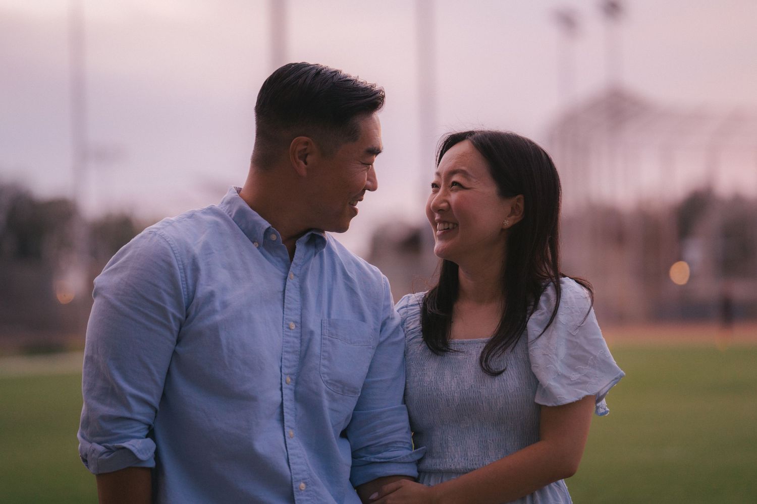 A smiling Asian couple gazes at each other affectionately outdoors at dusk on a green field.