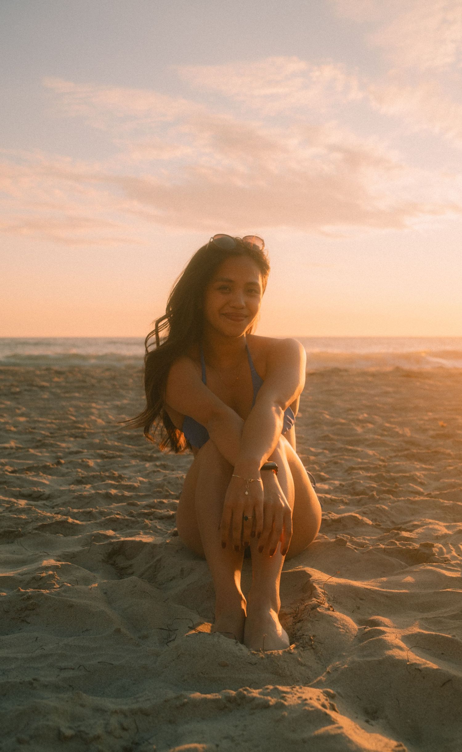Woman in blue bikini kneeling on beach sand during warm sunset glow.
