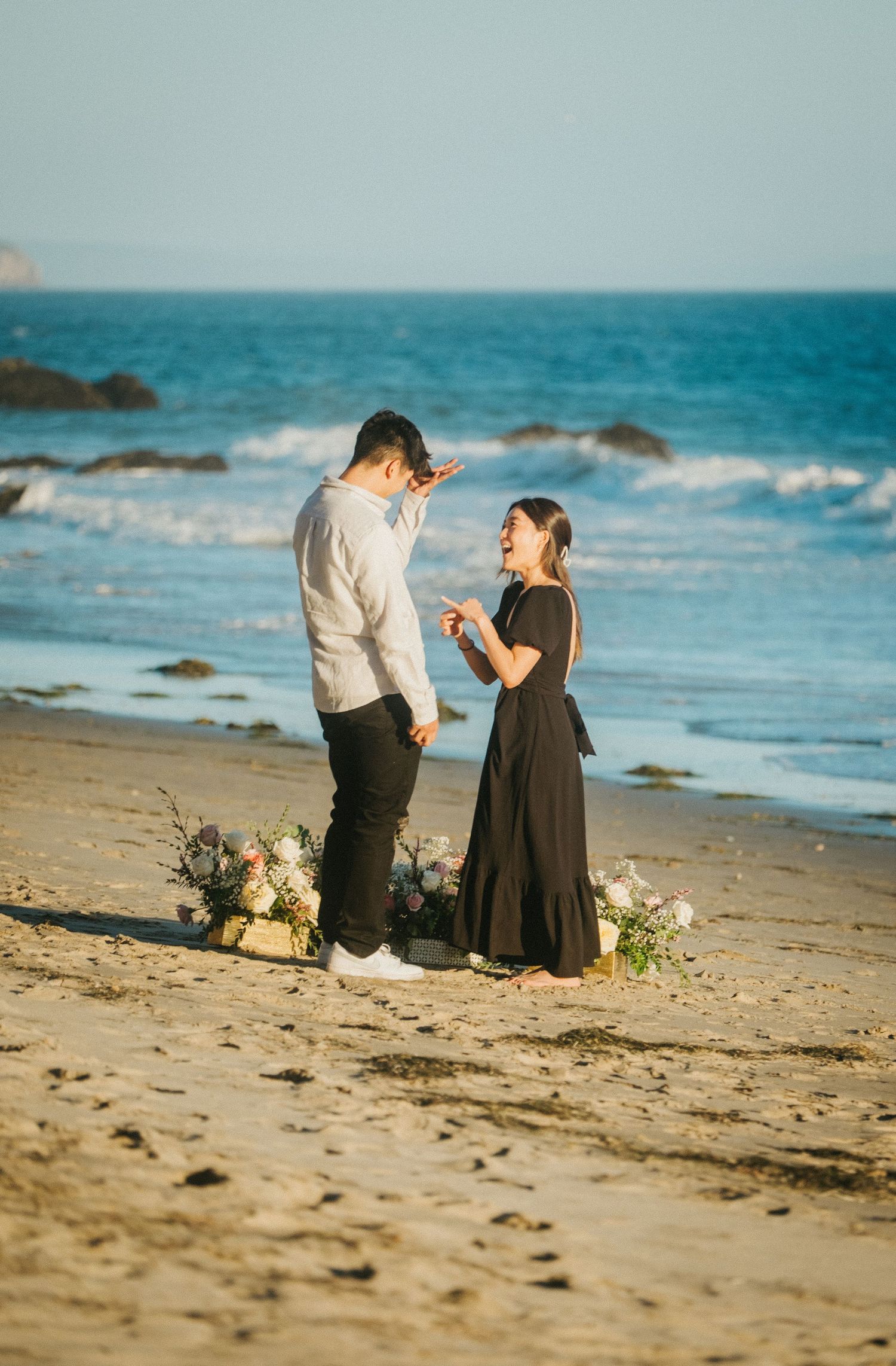 Couple sharing a joyful marriage proposal moment on a sandy beach with floral arrangements and ocean waves.