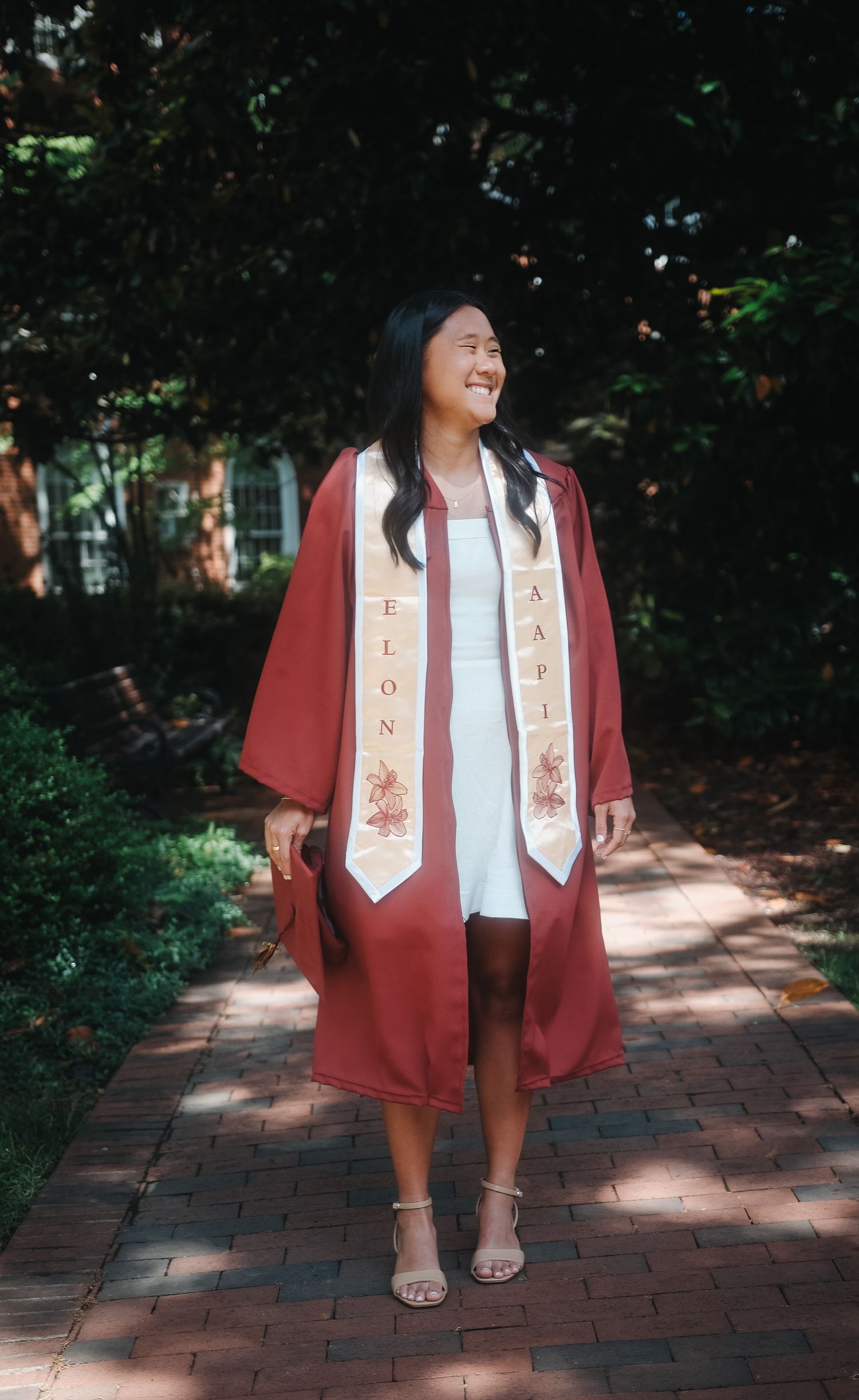 Joyful graduate smiles in maroon Elon University gown with AAPI stole on brick path.
