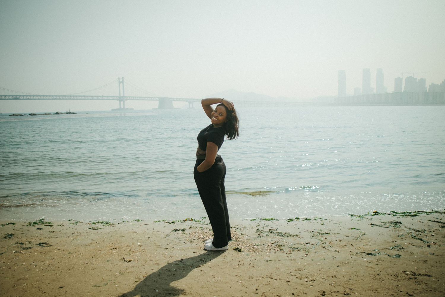 Woman in black outfit posing on a sandy beach with a suspension bridge and skyline behind her.