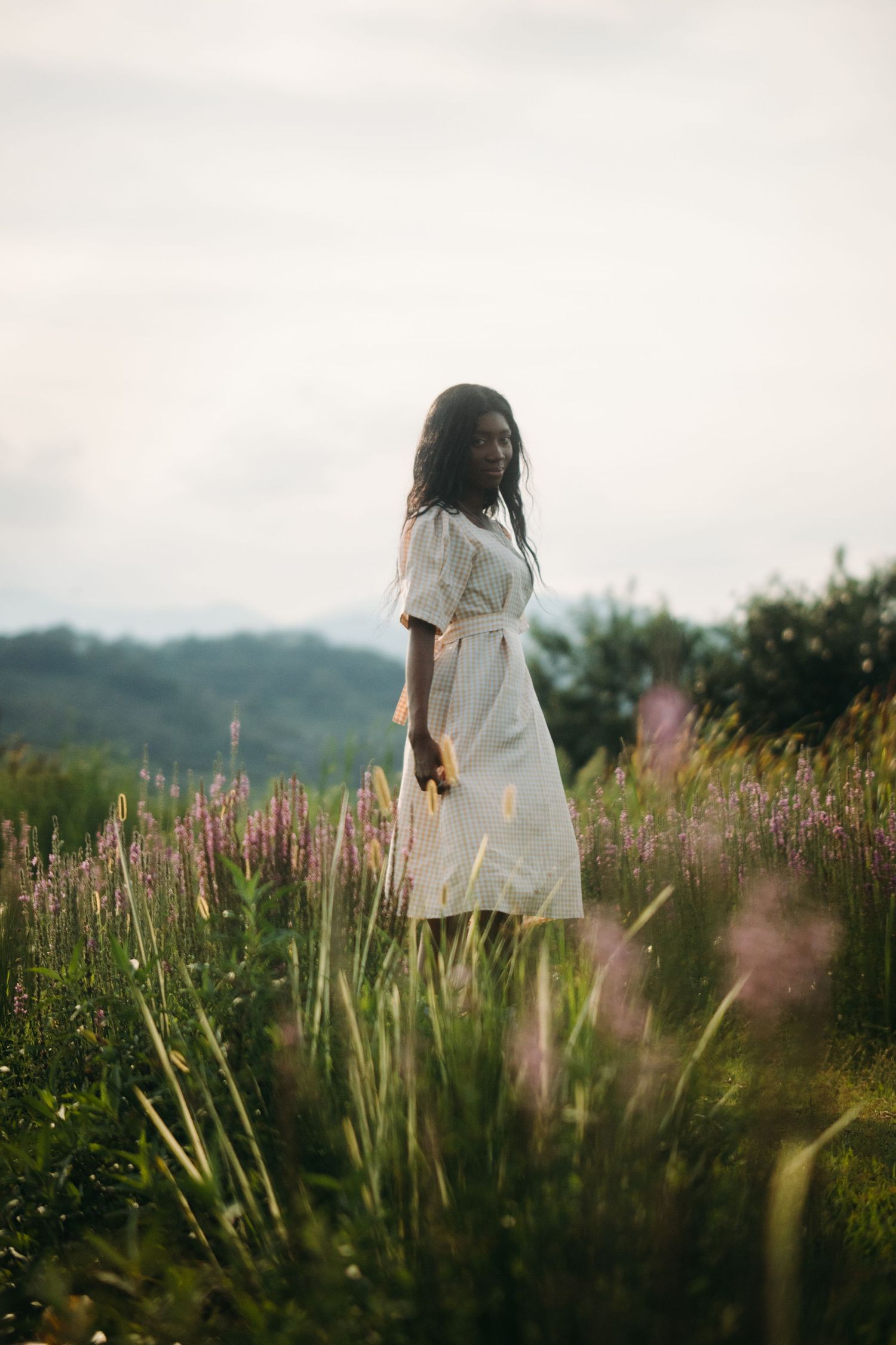 A young Black woman in a white midi dress stands gracefully among purple wildflowers in a lush green meadow.