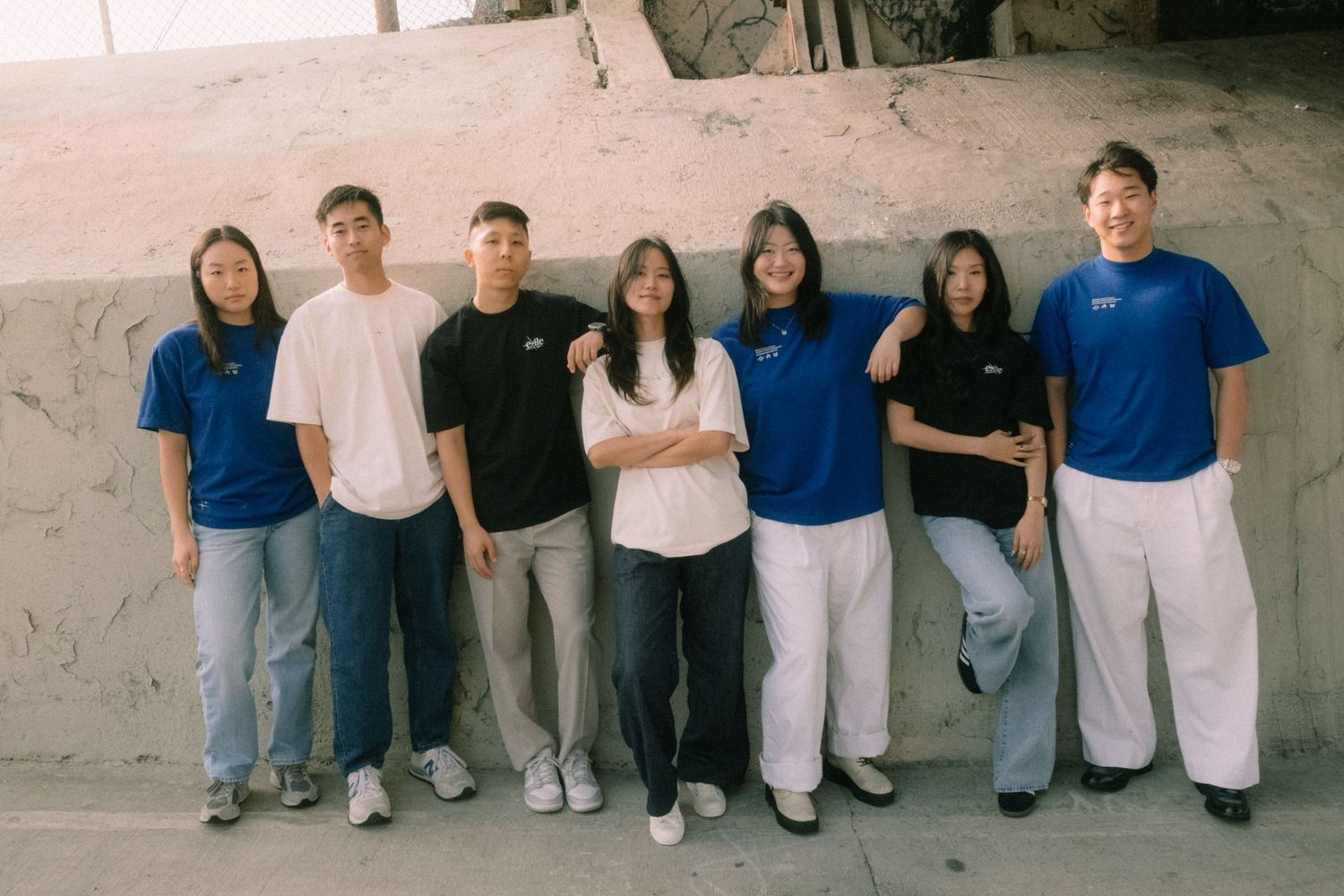 Seven young Asian people in matching blue shirts pose together outdoors against a stone wall.