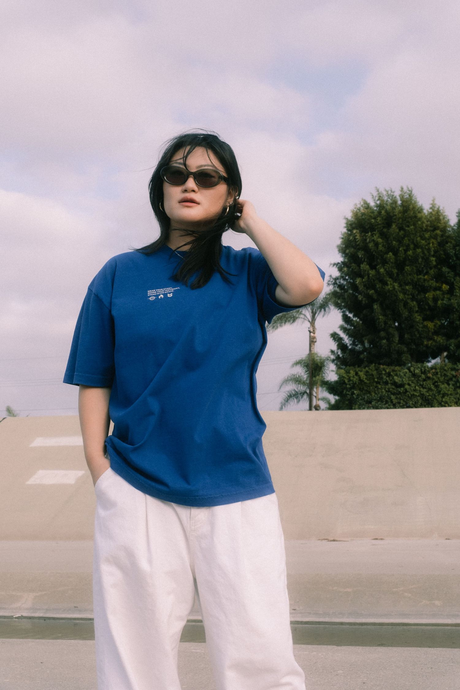 Woman in blue oversized tee, white wide-leg pants, and sunglasses posing outdoors under cloudy sky.