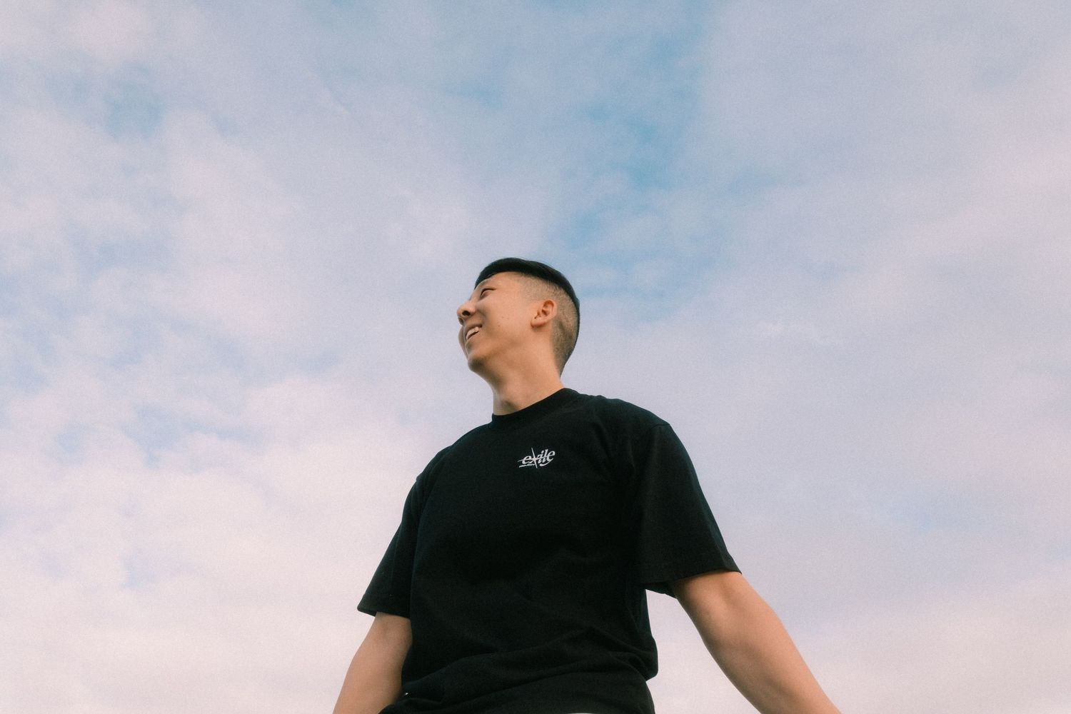 Young man in black t-shirt gazing upward against a cloudy blue sky, shot from low angle.