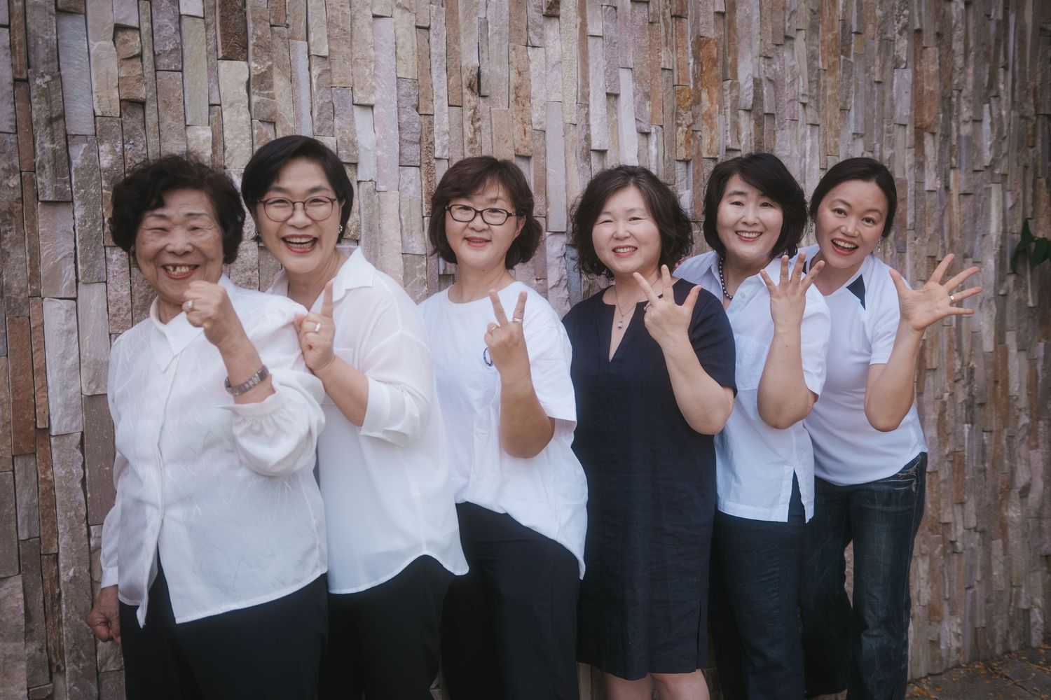 Six Asian women smiling and posing with peace signs against a rustic wooden wall backdrop.