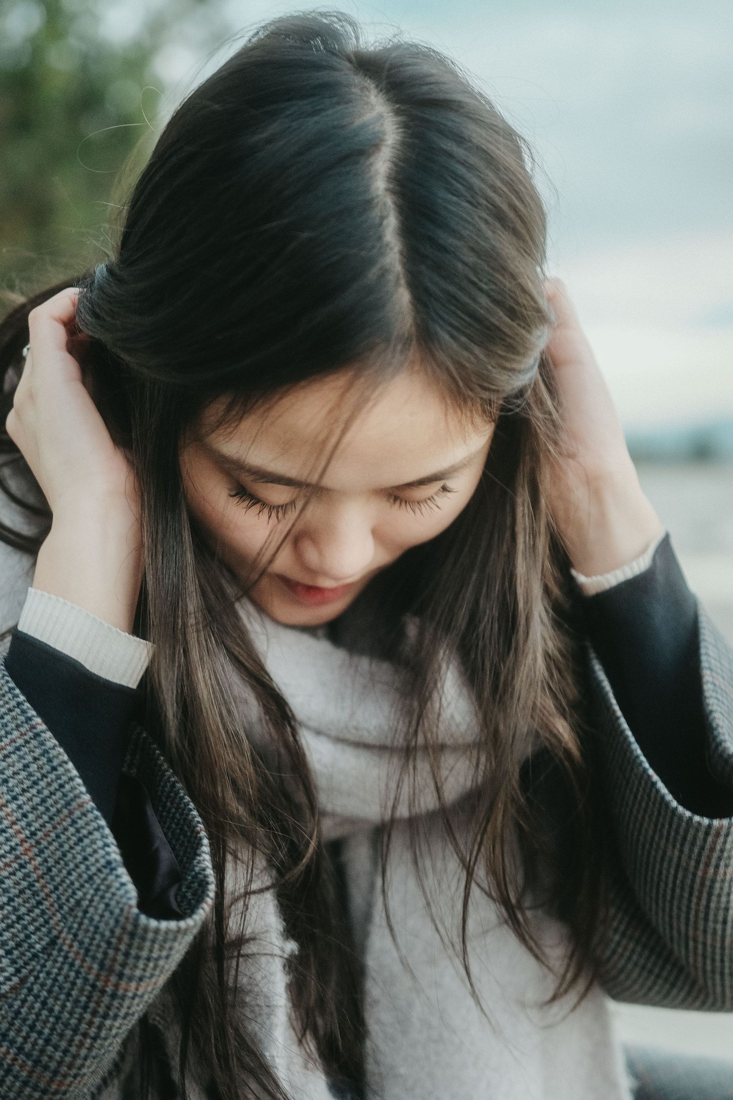 Young Asian woman with long dark hair holds her hair back with both hands, looking down pensively outdoors.