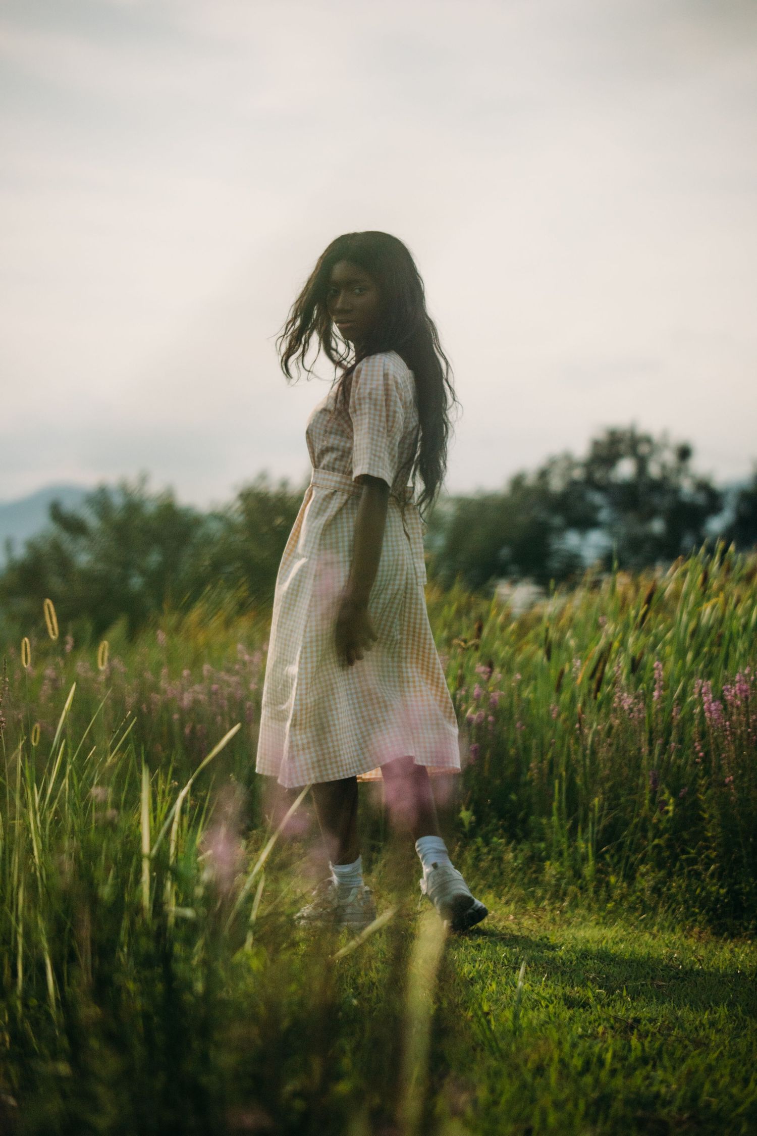 Young Black woman in striped dress walks through wildflower field with wind in her hair.