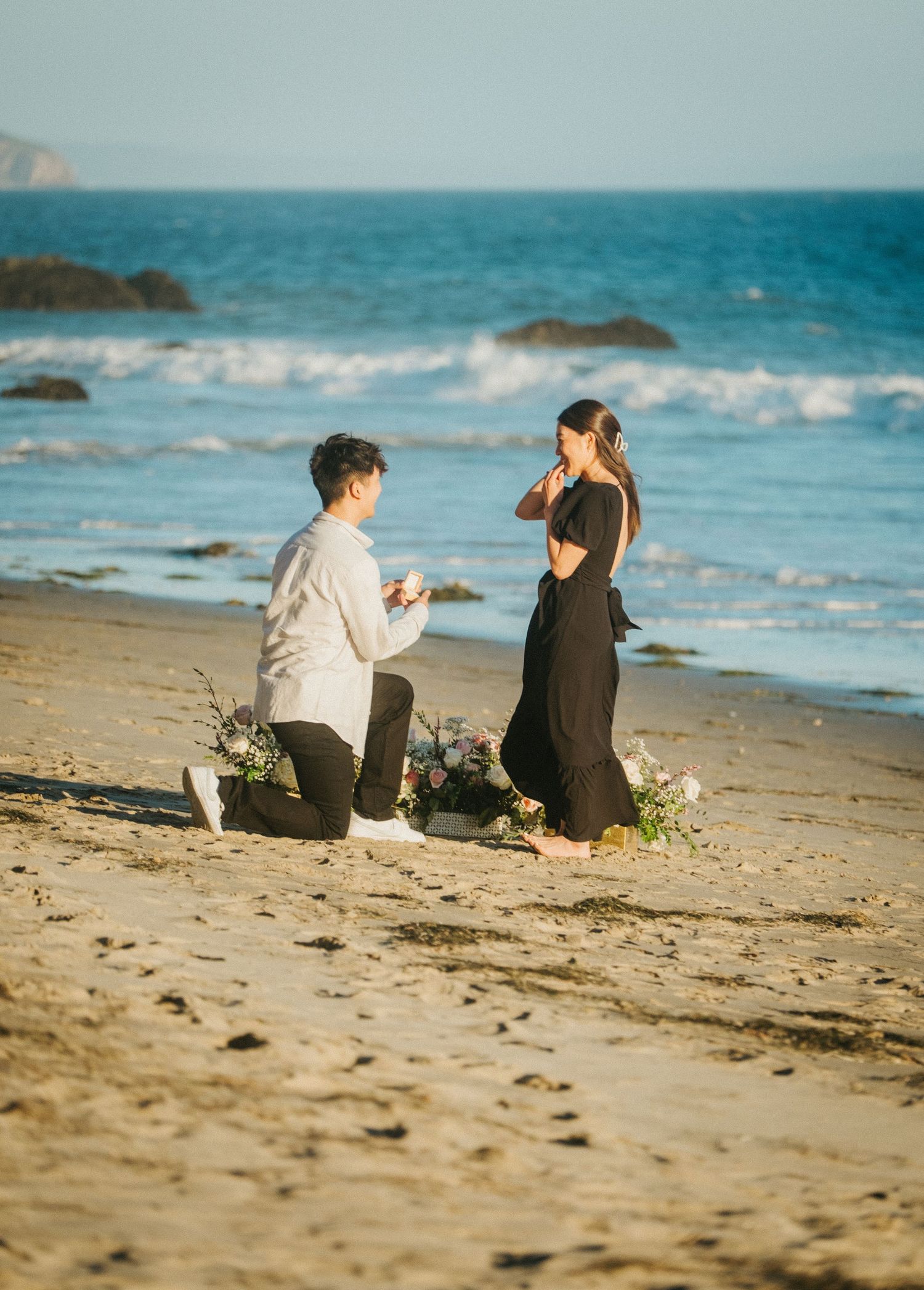 Man proposing on one knee to woman on sunny beach shoreline.