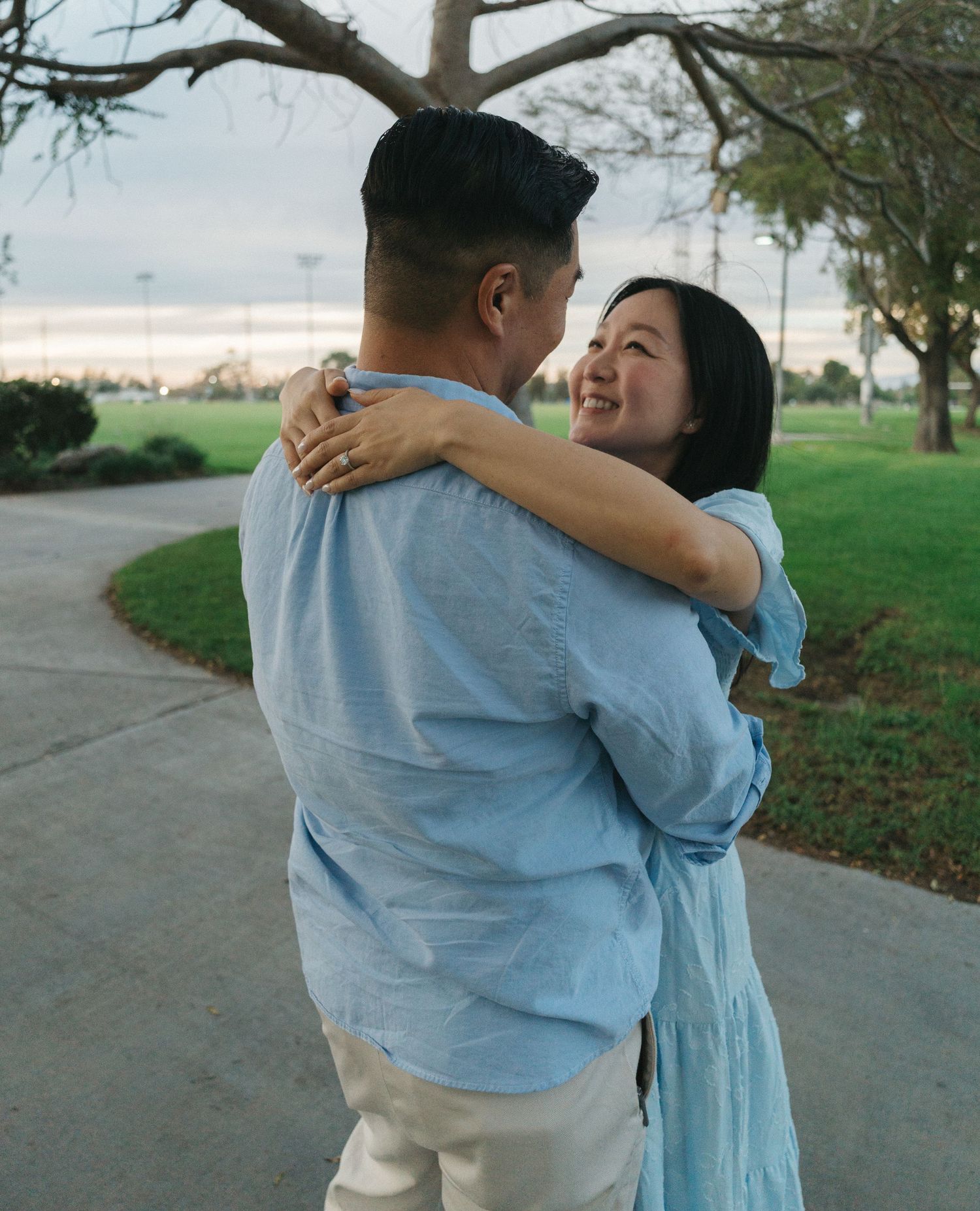 Smiling Asian couple embracing on park pathway at dusk, woman in blue dress hugging man.