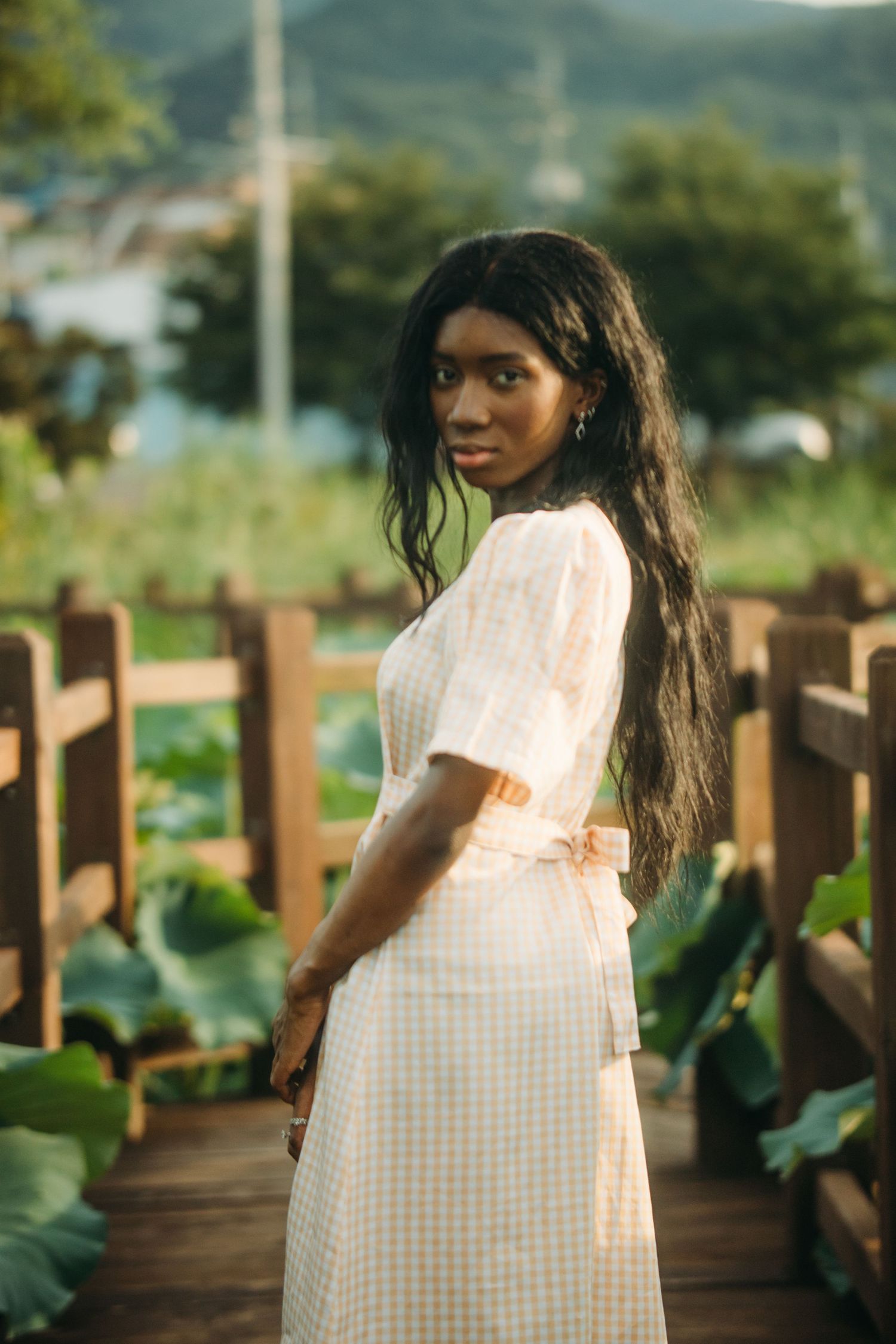 A young Black woman in a white gingham midi dress stands on a wooden path beside a garden.